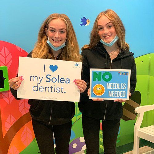 Two young women are holding signs that say `` i love my solea dentist '' and `` no needles needed ''.