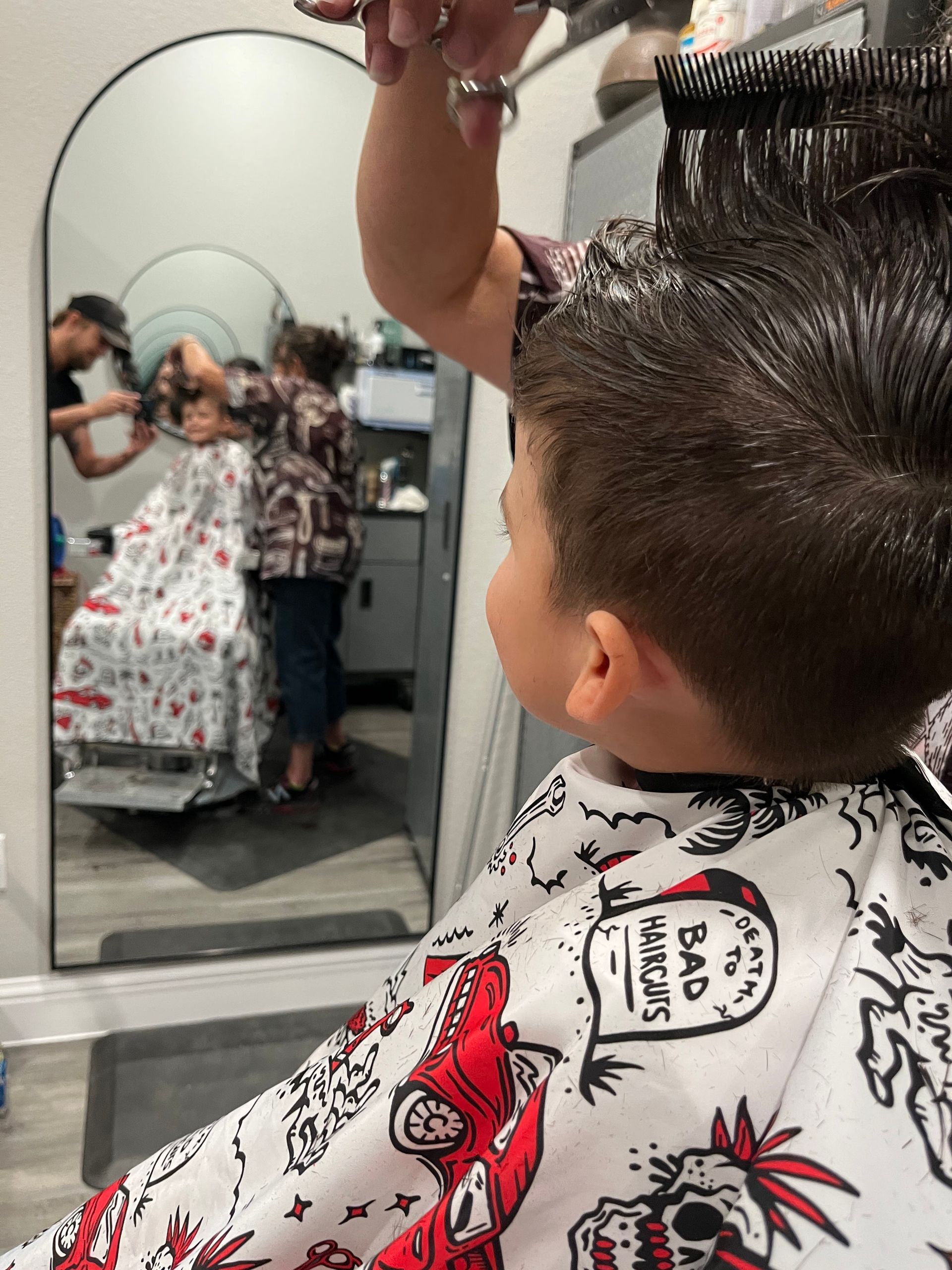 Child getting a haircut, seen from behind. Stylist is using scissors. The child is wearing a cape with skulls.