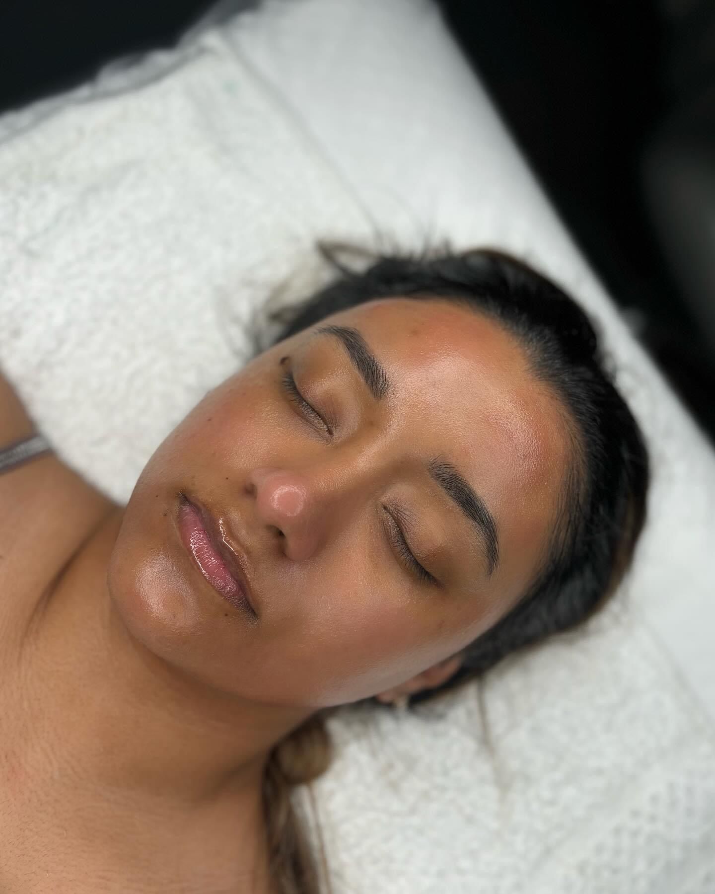 Woman with closed eyes, lying on a white surface, with a facial treatment. Her skin is glowing, and her lips are glossy.