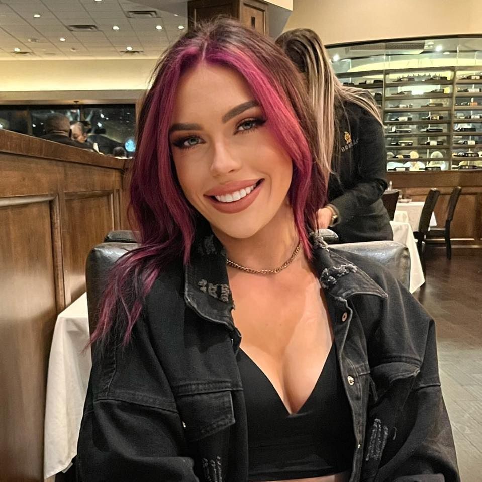 Woman with pink-streaked hair smiles, wearing a black jacket and necklace, at a restaurant table.