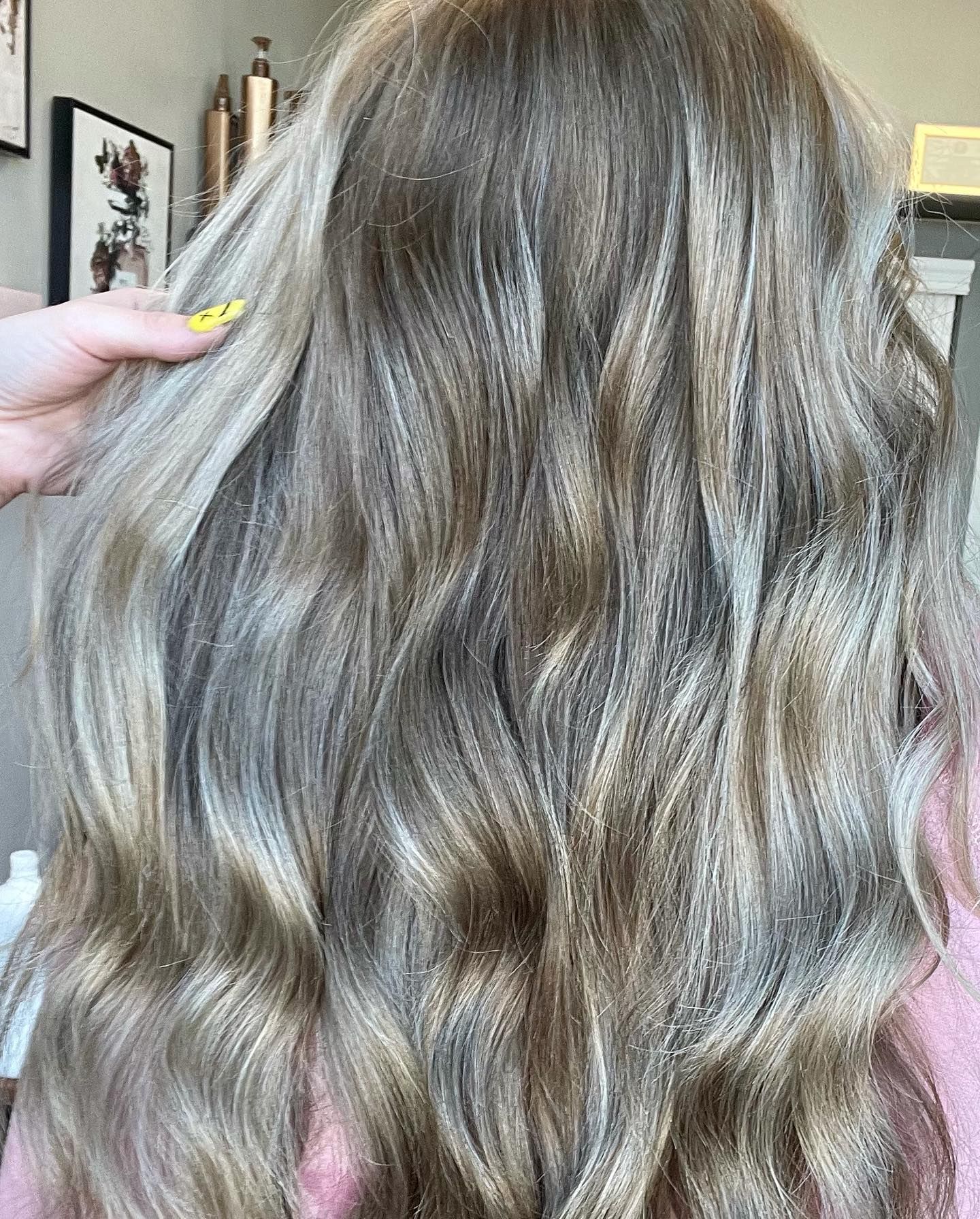 Woman's wavy, highlighted blonde hair. A hand with a yellow painted nail touches the hair.