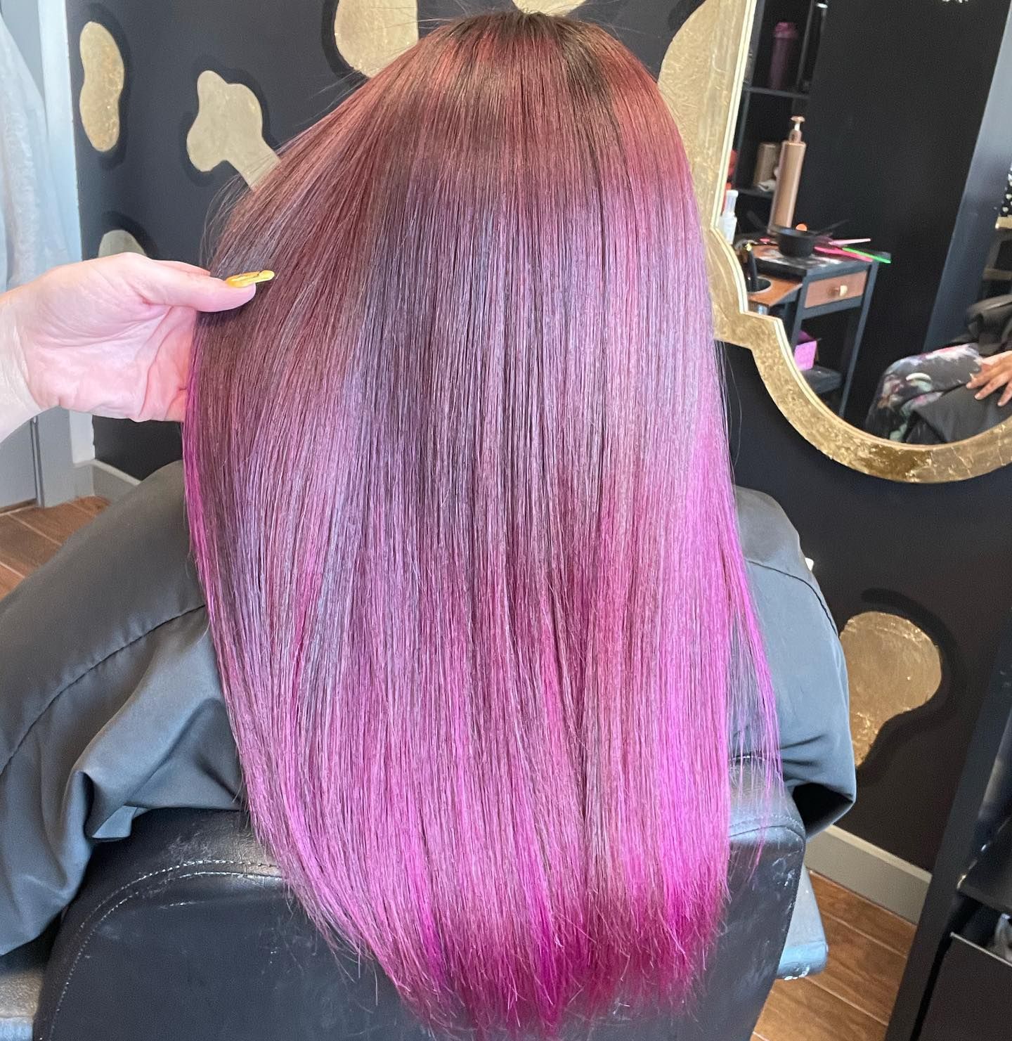 Woman's long hair, top dark red fading to vibrant pink. A hand is smoothing the hair in a salon setting.