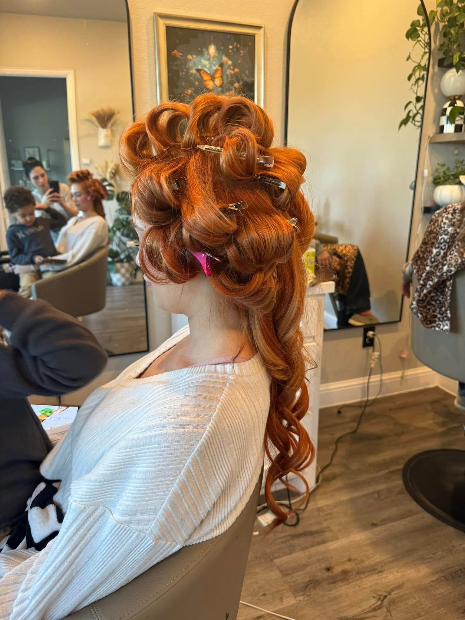Woman with bright red hair in rollers at a salon, seated and facing away. Other people and a mirror are visible.