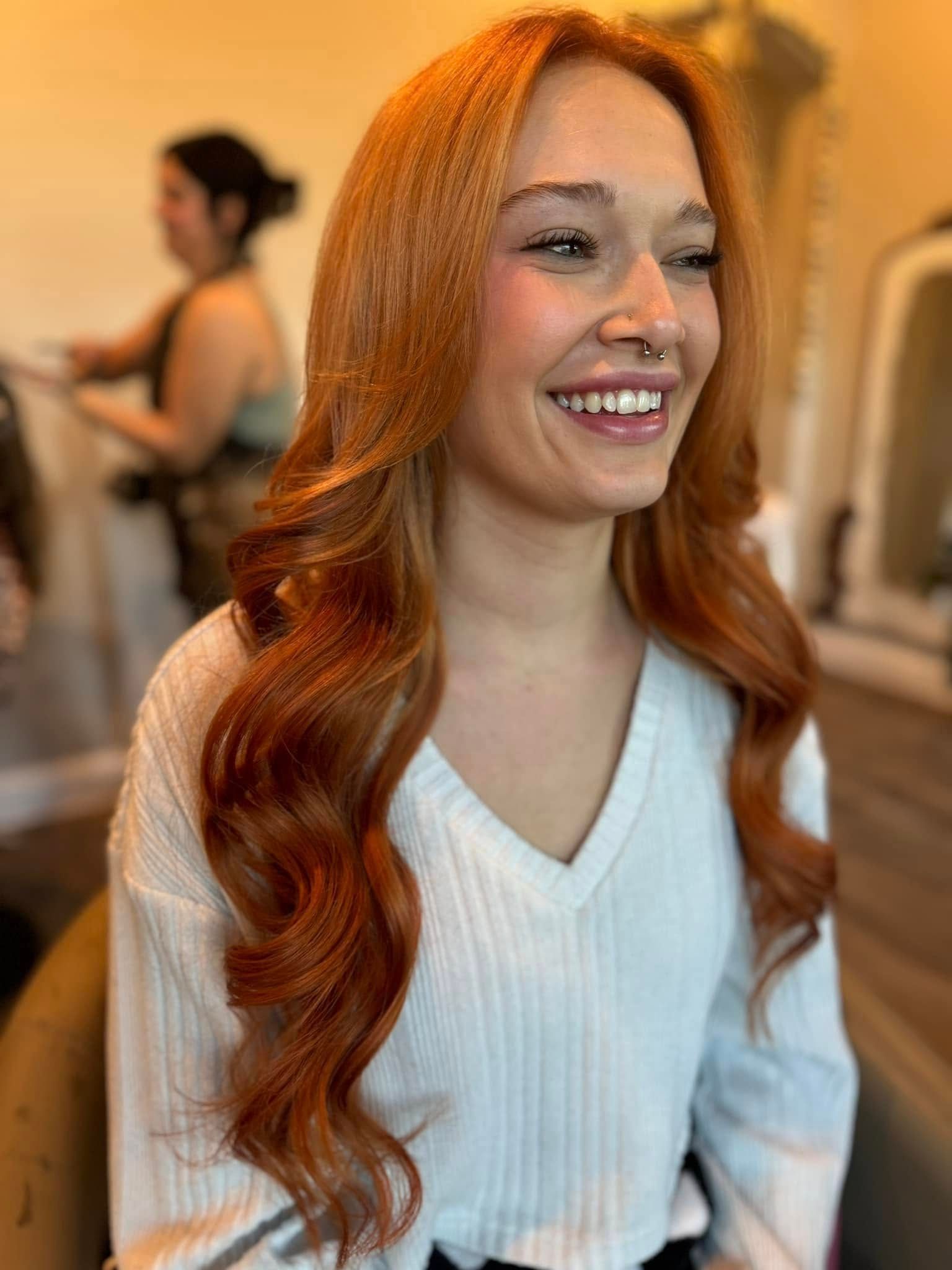 Woman with long, wavy, red hair smiles, wearing a white sweater, in a salon setting.