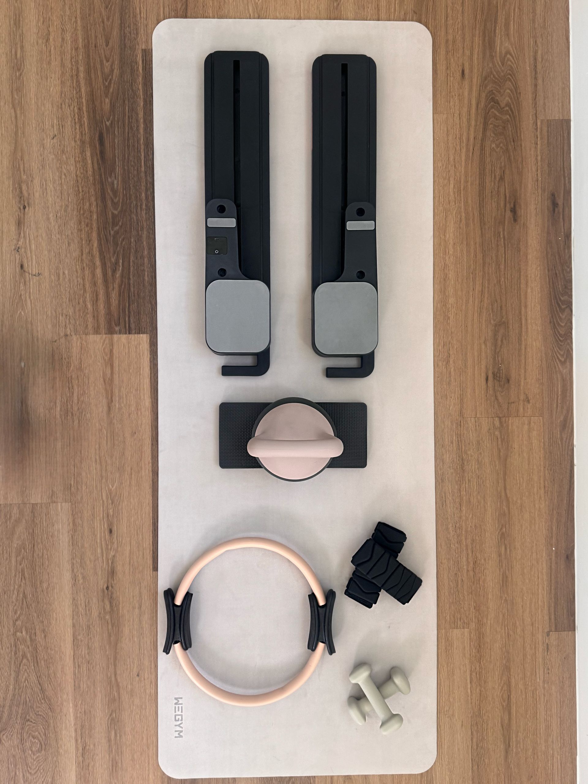Pilates equipment: two black leg attachments, a central bar with pink cushion, a ring, weights, and grips on a light mat.