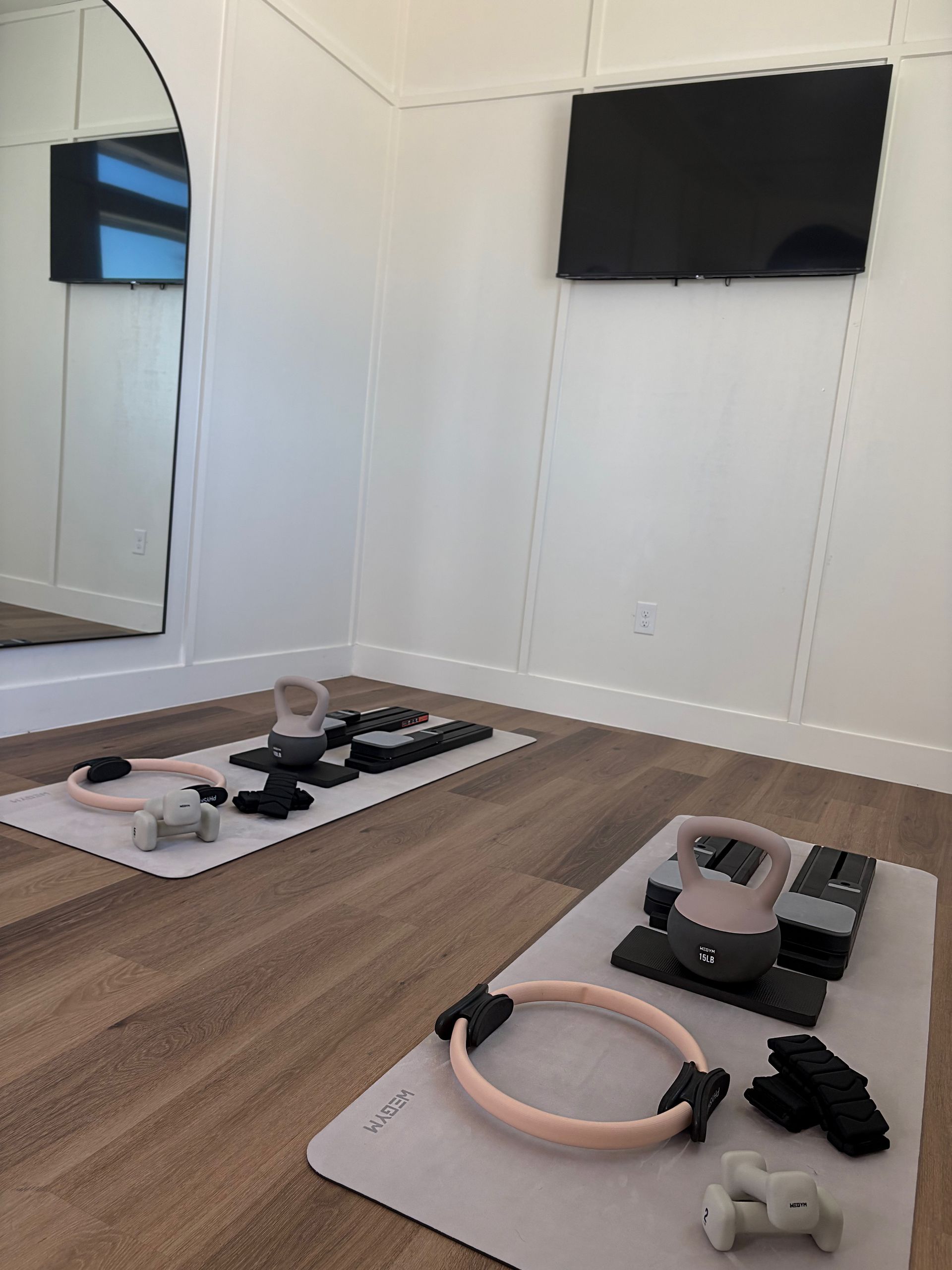 Workout room with exercise equipment on mats: pink rings, kettlebells, weights, and TV on wall.