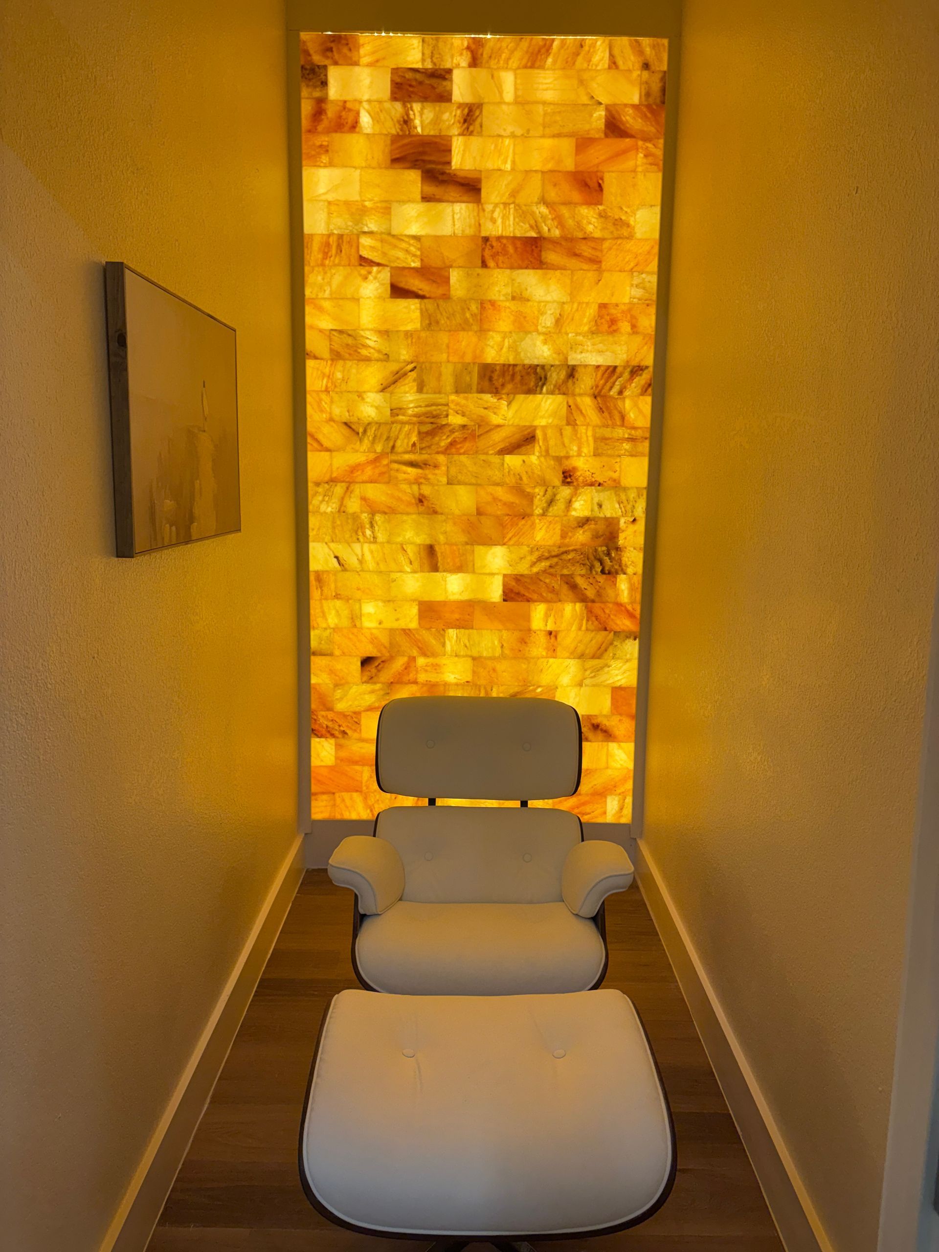 White chair and ottoman in a narrow space with glowing, yellow-lit brick wall behind it. A painting hangs on the side wall.