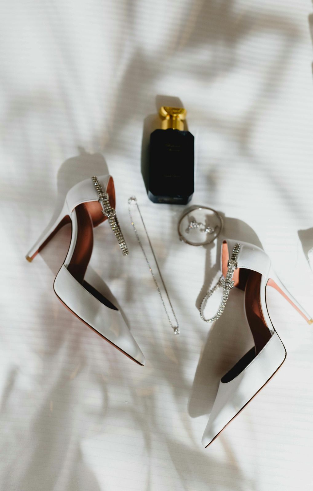 White high heels, black perfume bottle, jewelry on white fabric.