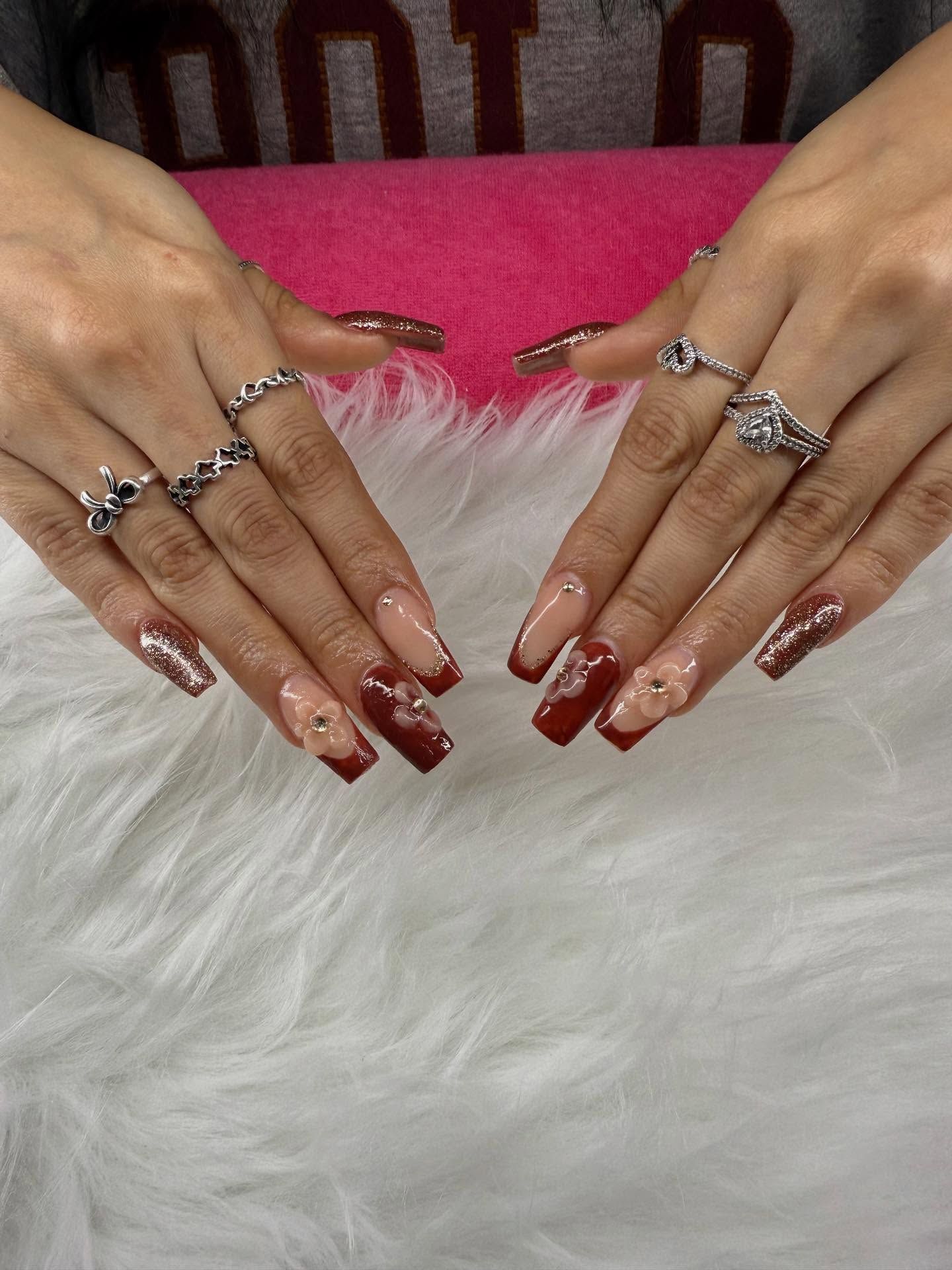 Hands with long, burgundy and nude-colored acrylic nails adorned with rhinestones, plus silver rings.