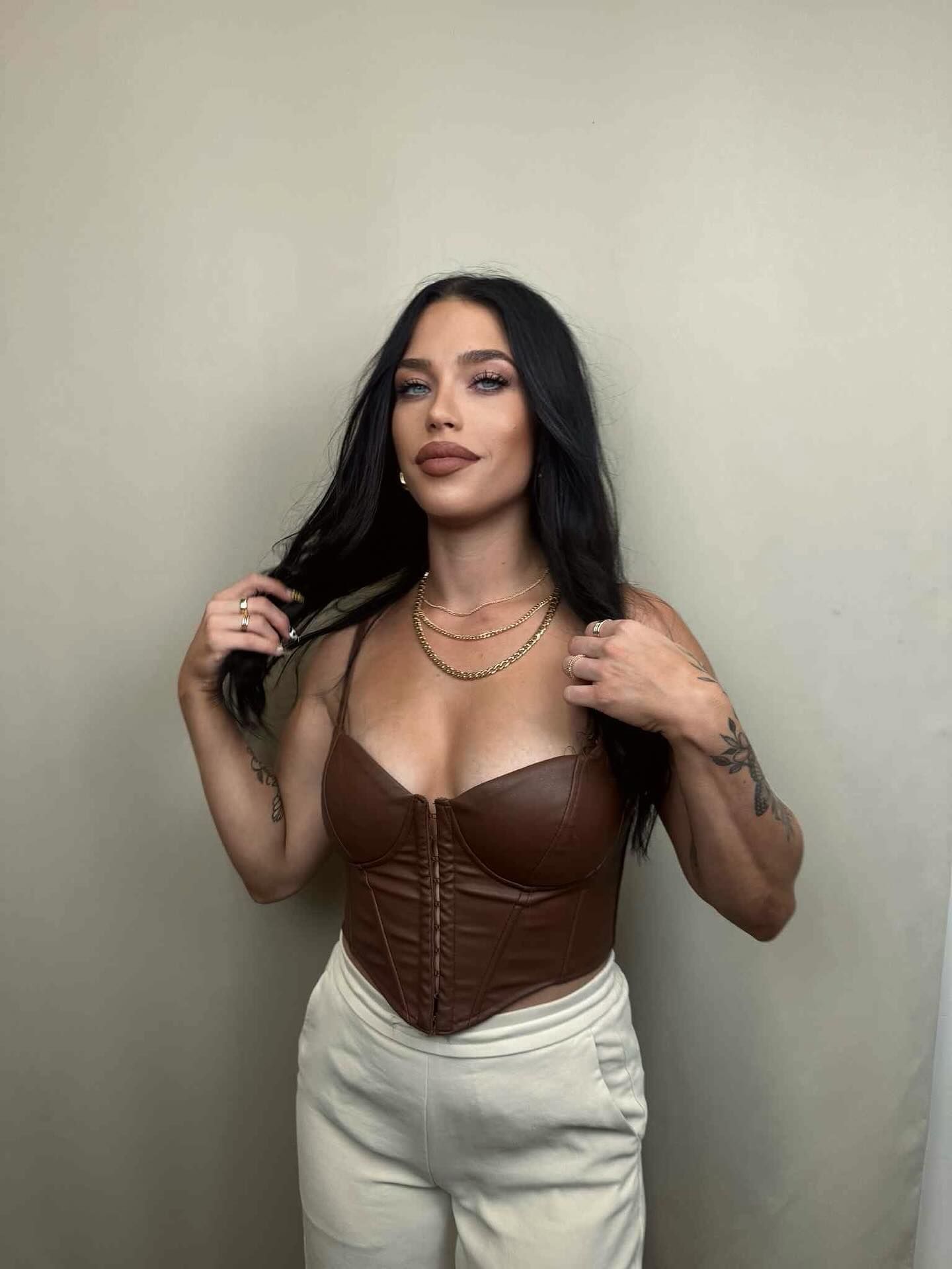 Woman in brown corset top and cream pants, touching hair.