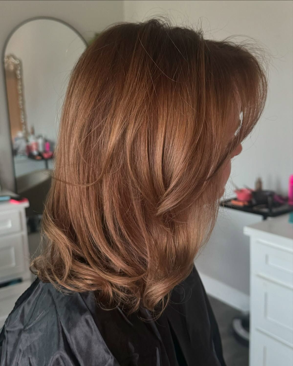 Woman with copper-colored layered hair, side view. In a salon, a light-colored mirror and furniture are visible.