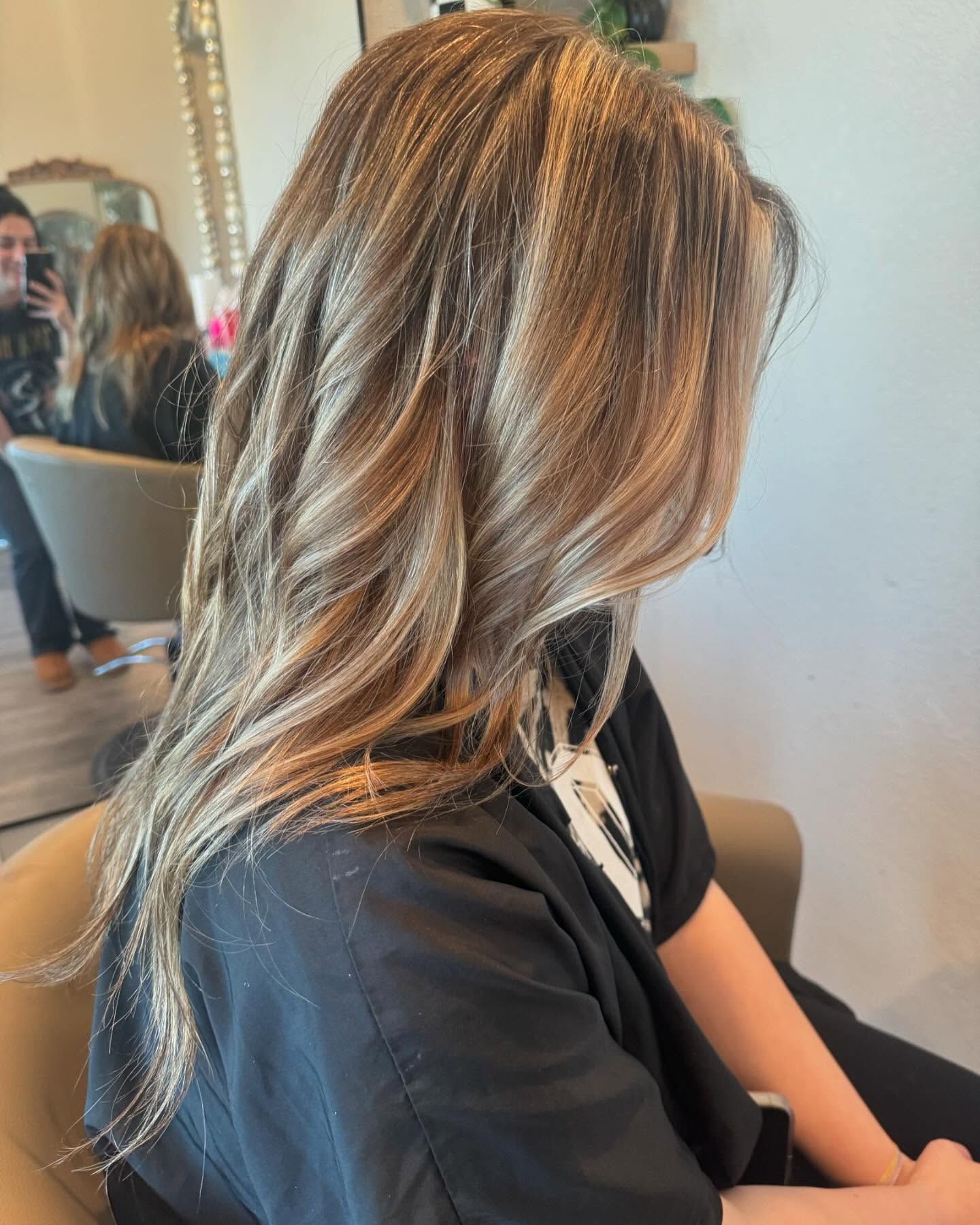 Woman with long, wavy, highlighted hair; seated in a salon chair. Highlights are blonde/light brown.