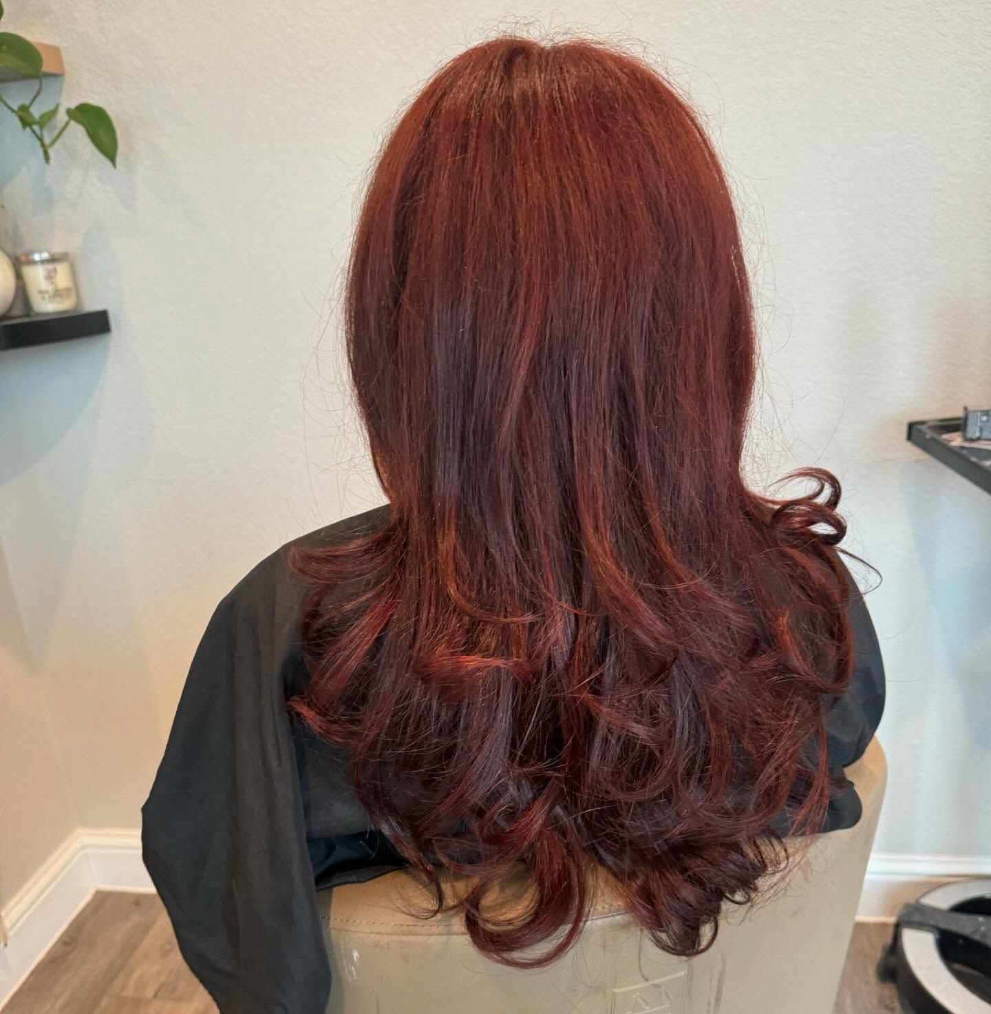 Woman with long, wavy auburn hair. Sitting in a chair in a salon, wearing a black cape.