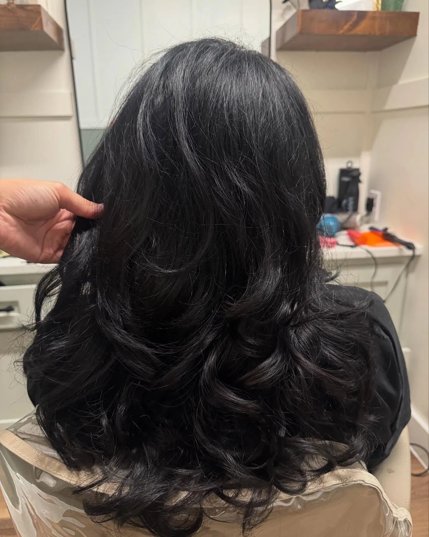 Person with long, black wavy hair sitting in a salon chair, hand touching the hair.
