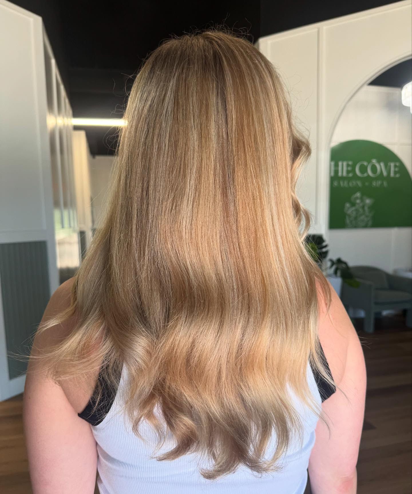 Woman with light brown hair, standing inside a building. The hair is long and wavy. The woman wears a white tank top.