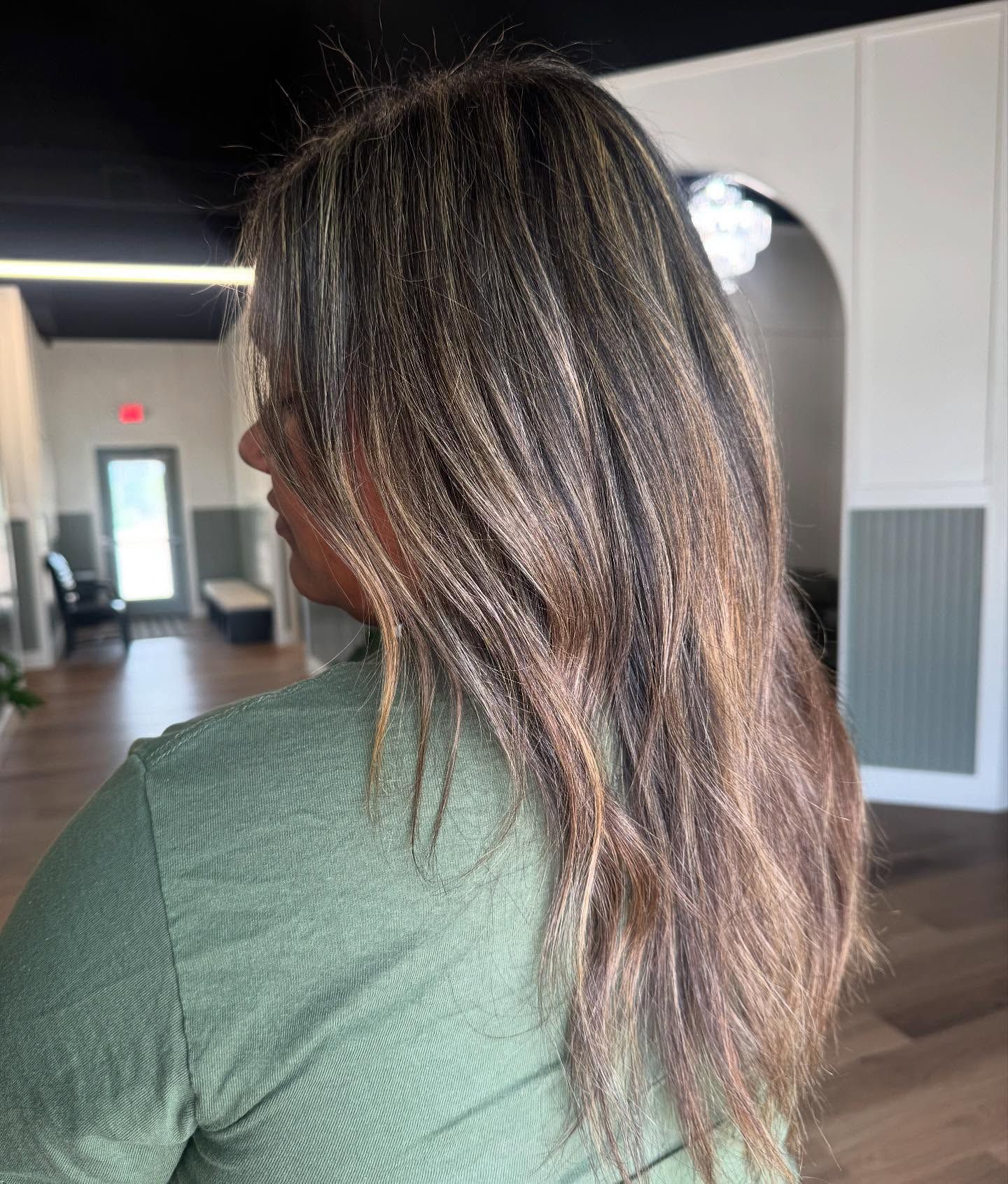 Woman with long, layered brown hair, some blonde highlights. Stands in a salon, wearing green top.