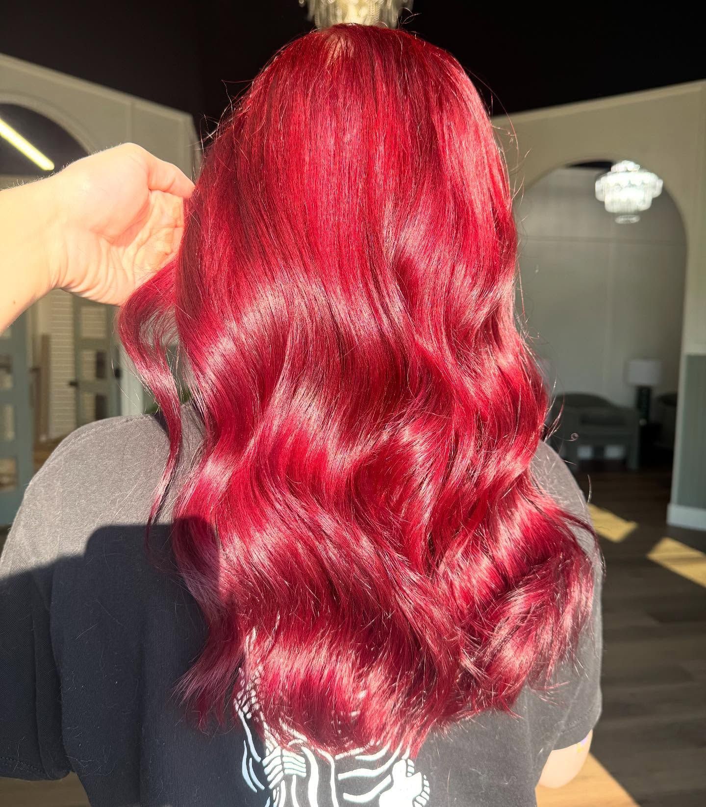 Woman with long, wavy, bright red hair in a salon setting. A hand gently touches the hair.