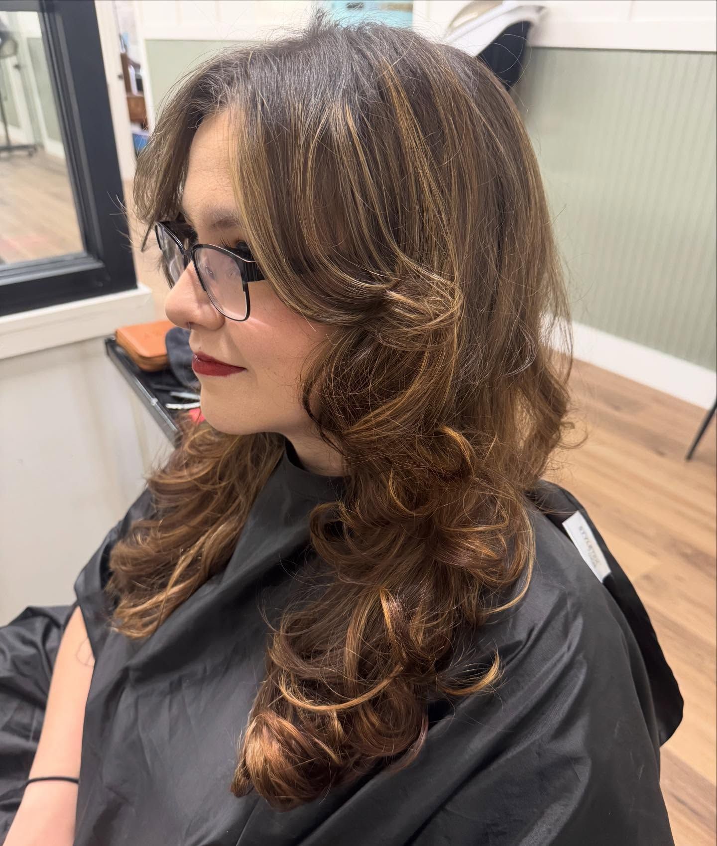 Woman with long, wavy brown hair with highlights, wearing glasses and a black salon cape.