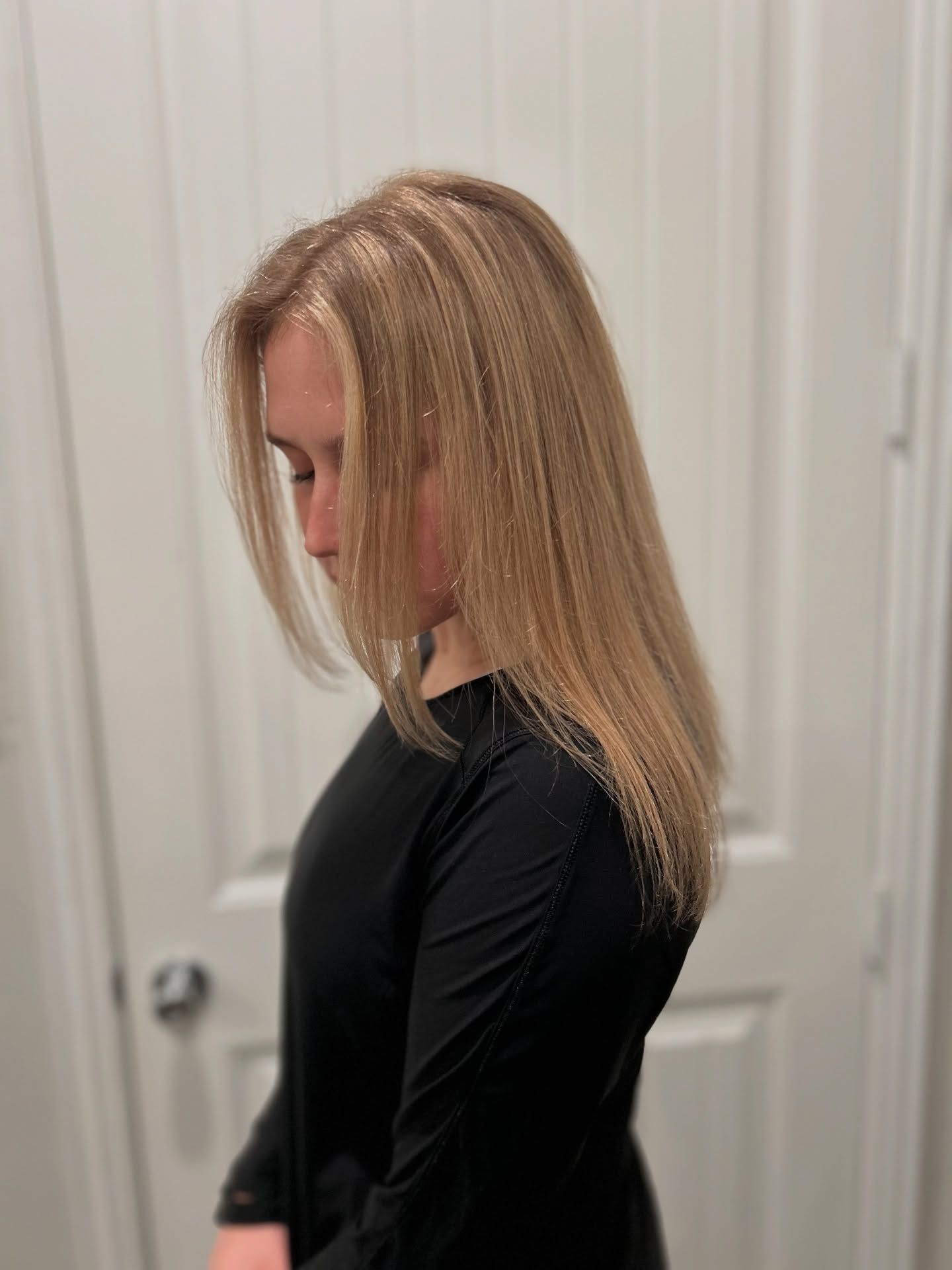 Woman with long, light brown hair wearing a black long-sleeved shirt, standing in front of a white door.