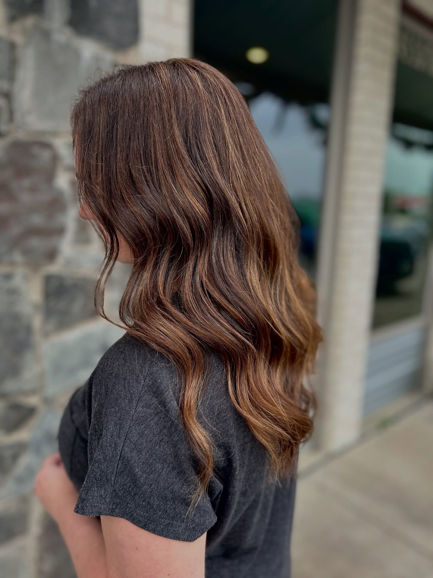 Woman with long, wavy brown hair, wearing a gray shirt, standing outdoors near a stone wall.