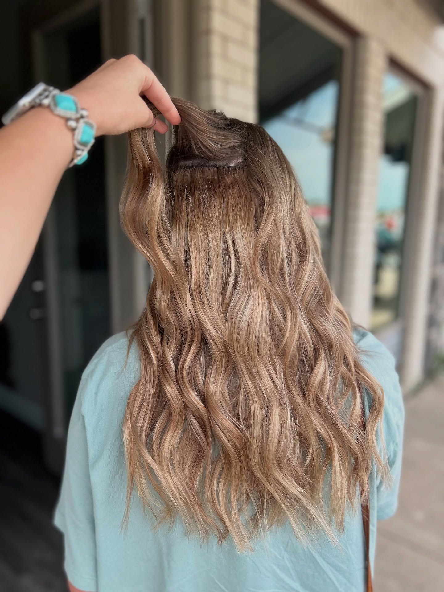 Person with long, wavy blonde hair; hand holding a section. Light blue top, turquoise jewelry, outdoors.