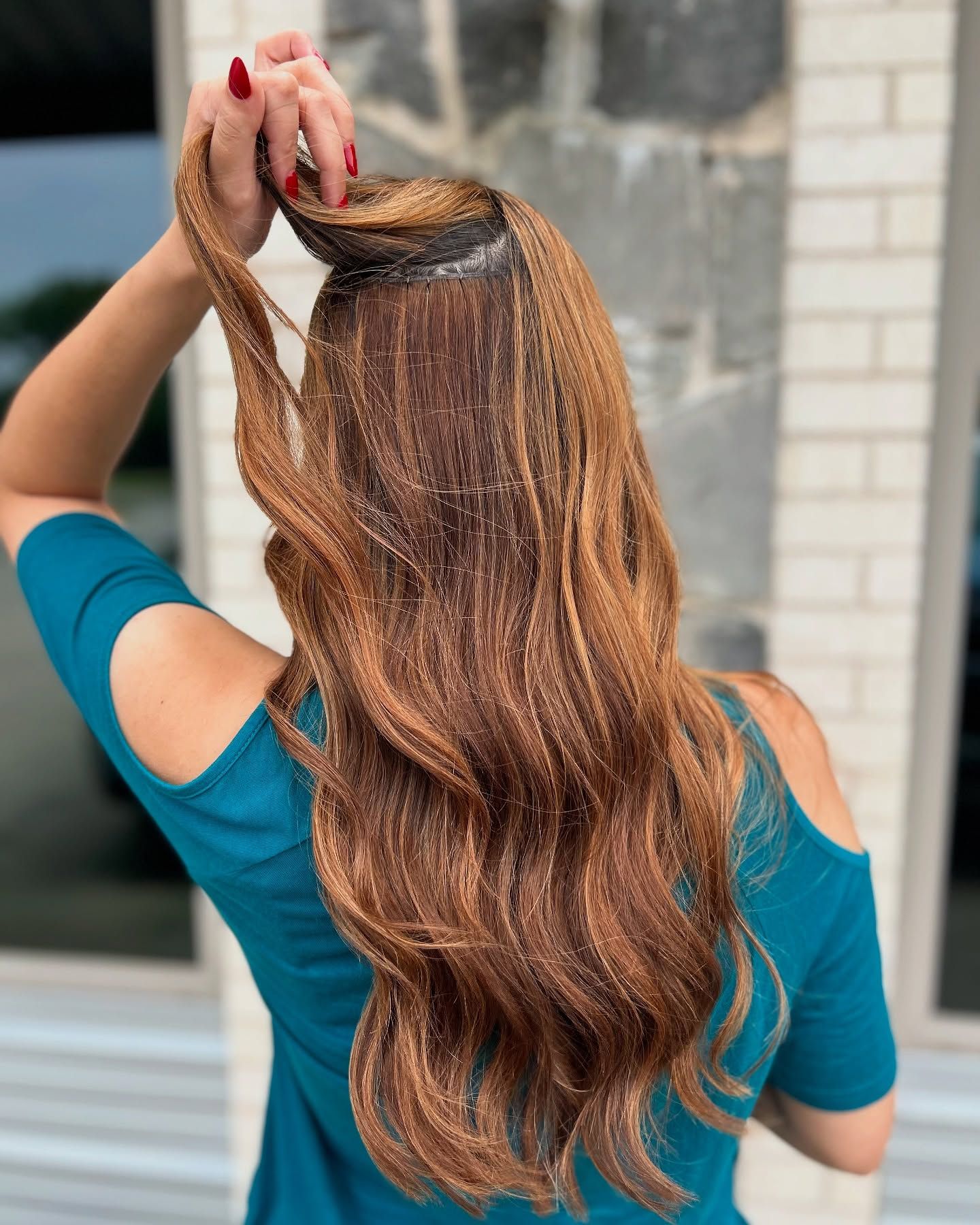 Woman with long, wavy, auburn hair, wearing a teal top, holding up a section of hair.