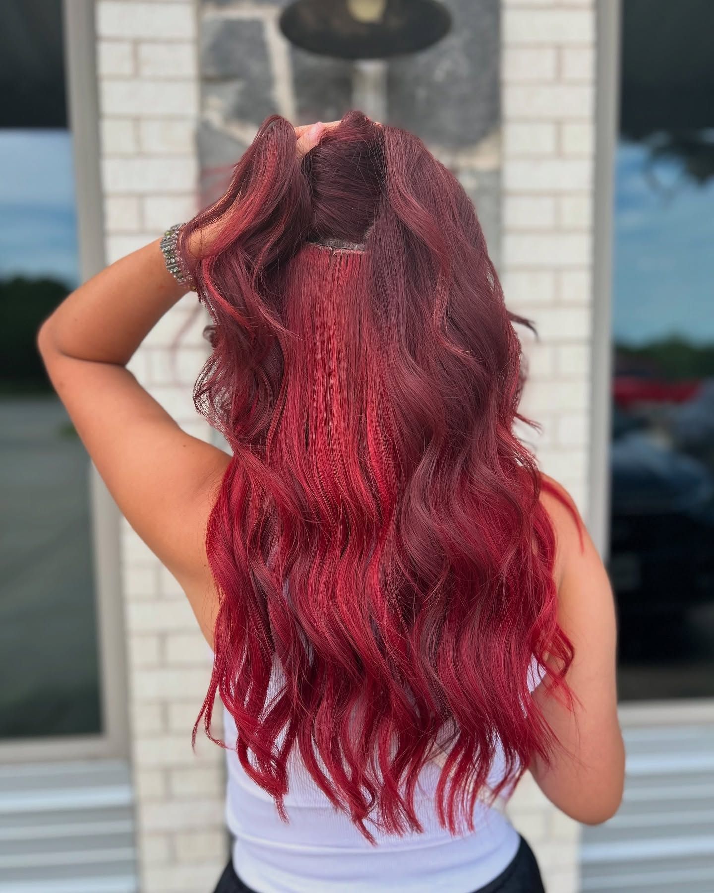 Woman with long, red wavy hair, standing in front of a white brick wall, arms raised, showing back.