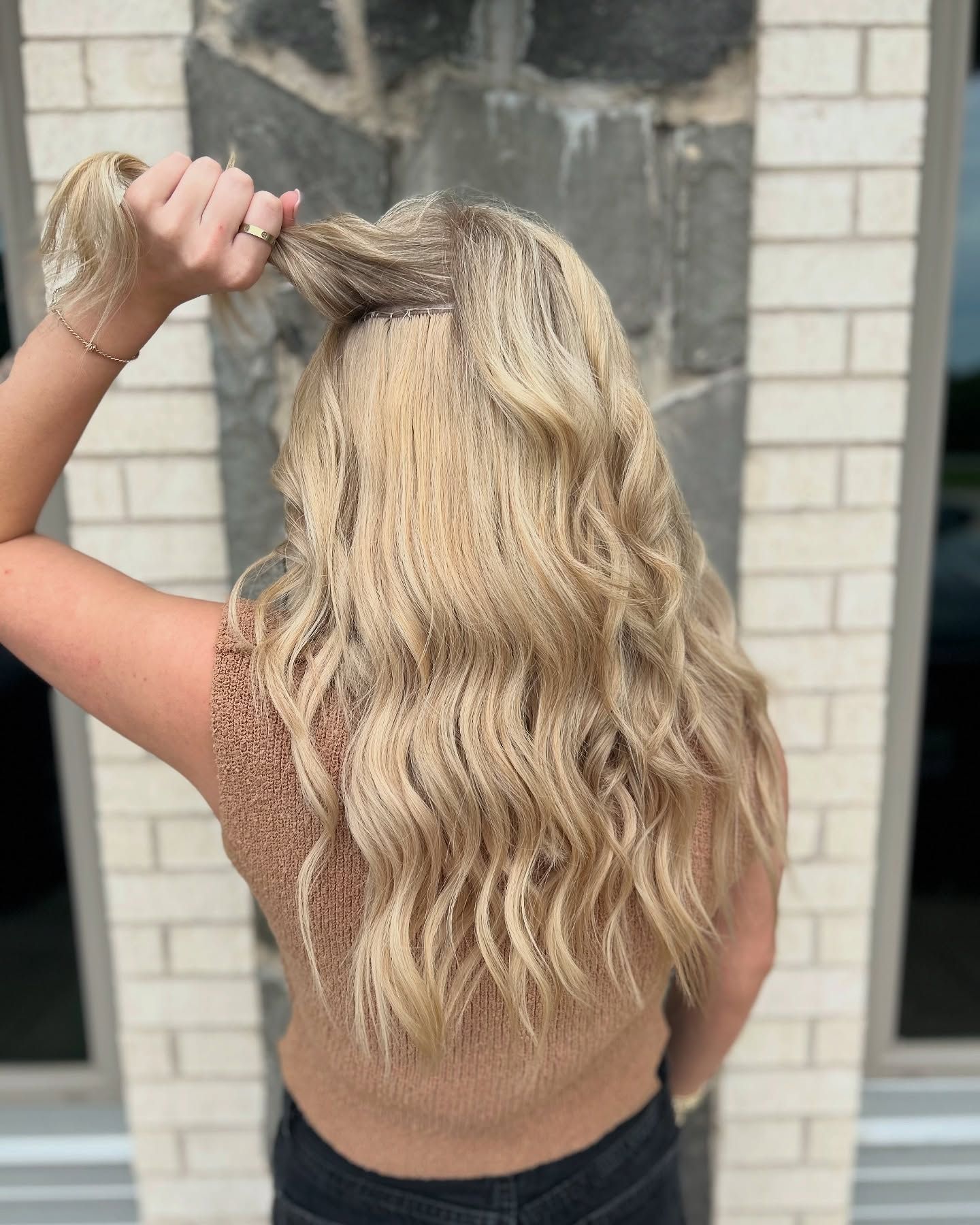 Woman with long, wavy blonde hair holding some strands up, wearing a tan top. Brick and stone background.