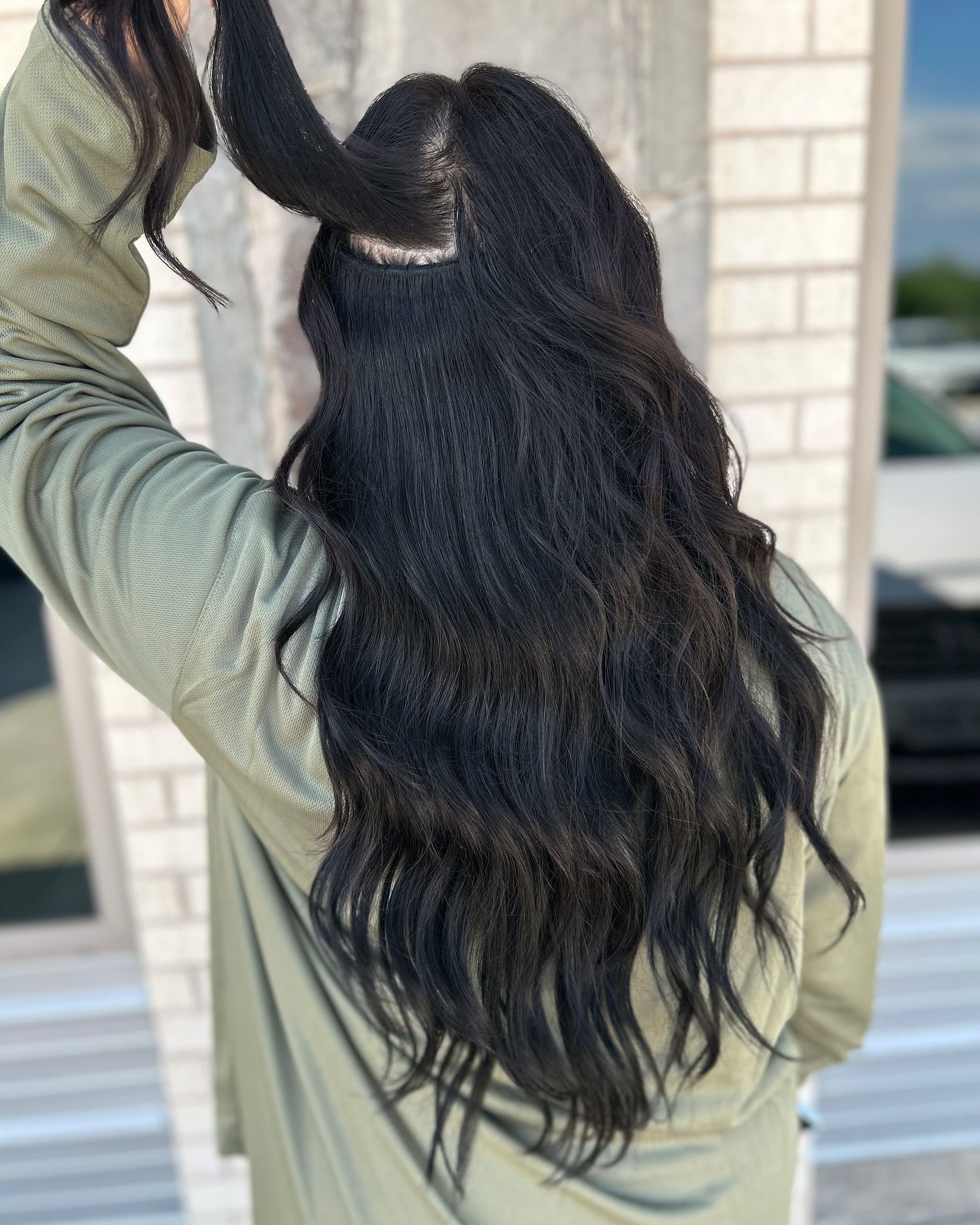 Woman with long black wavy hair, wearing a green shirt, holding up hair near a brick wall.