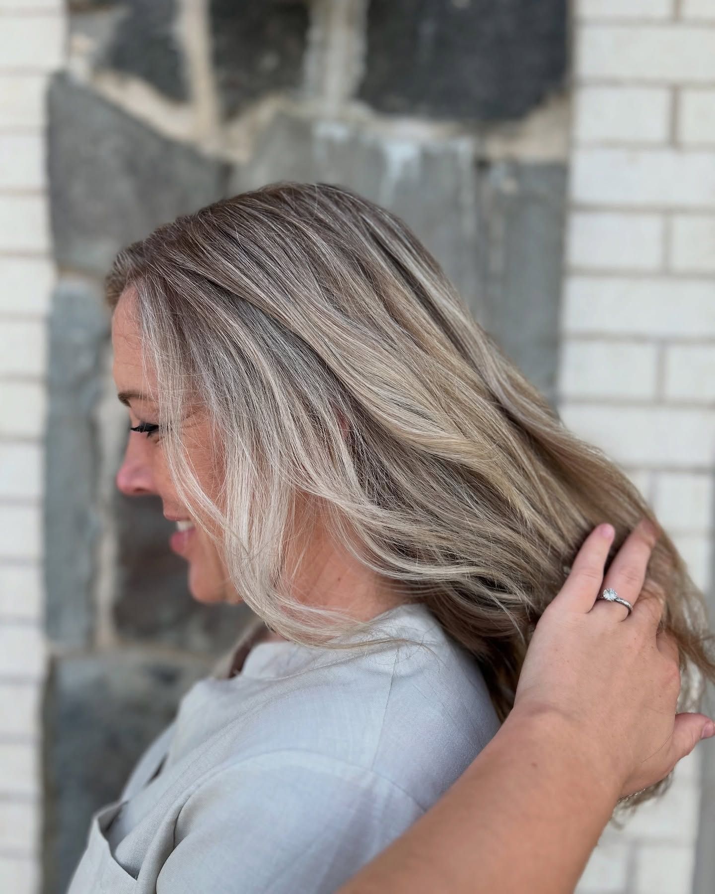 Woman with blonde hair smiles, hand in hair, against a stone wall.