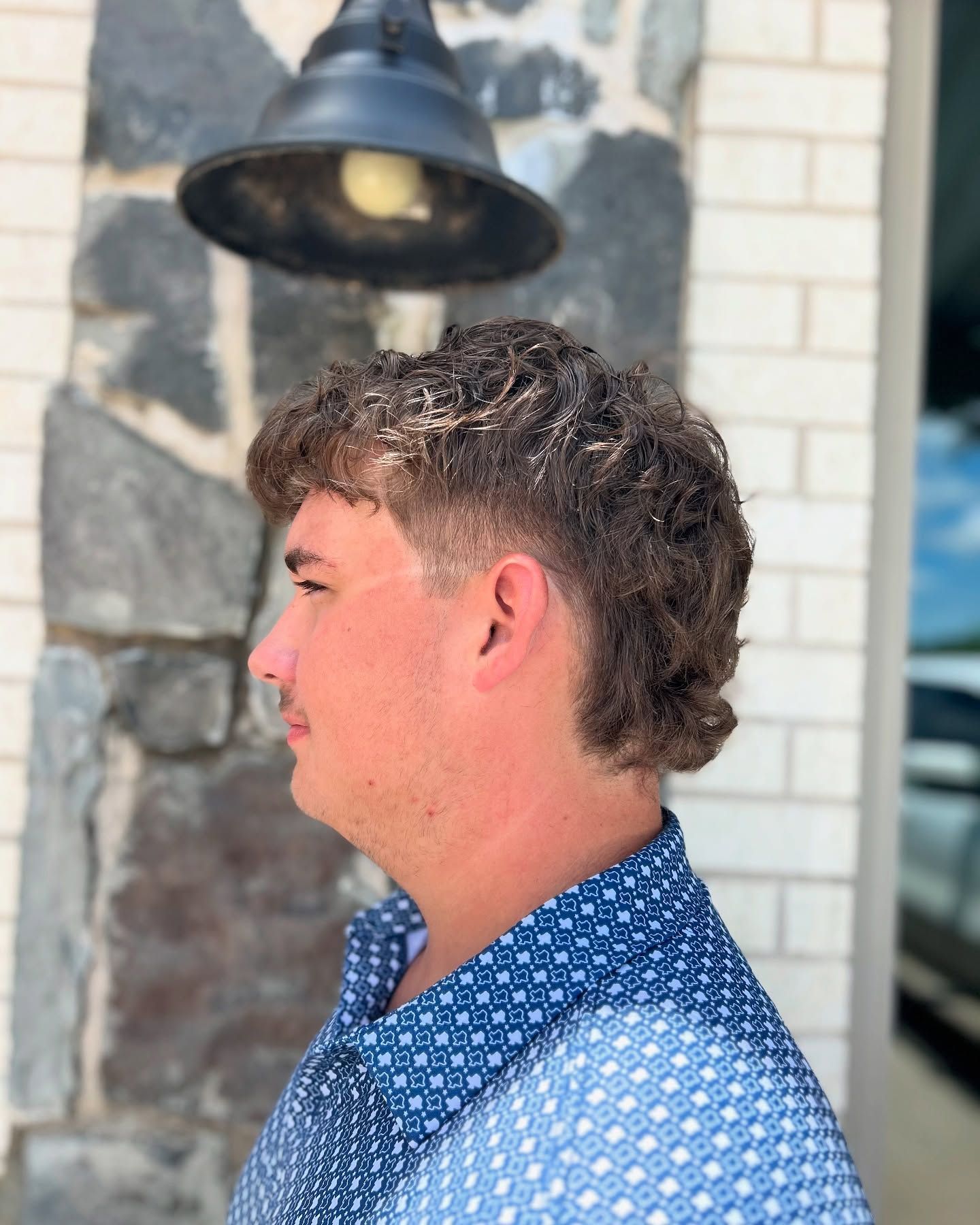 Man with curly hair and a mullet hairstyle, wearing a blue patterned shirt, outdoors.