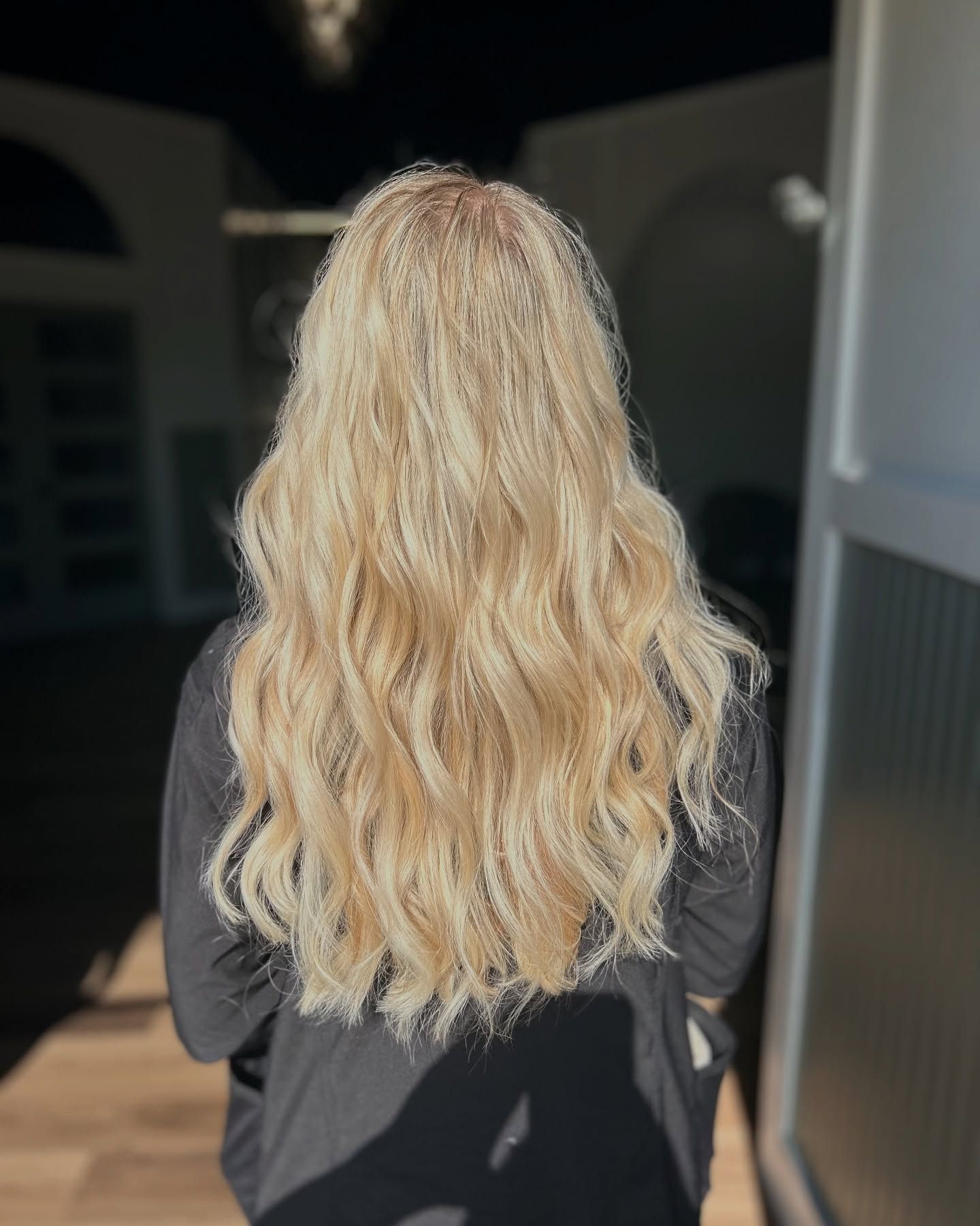 Blonde wavy hair cascading down back of person wearing a dark top, standing near a bright doorway.