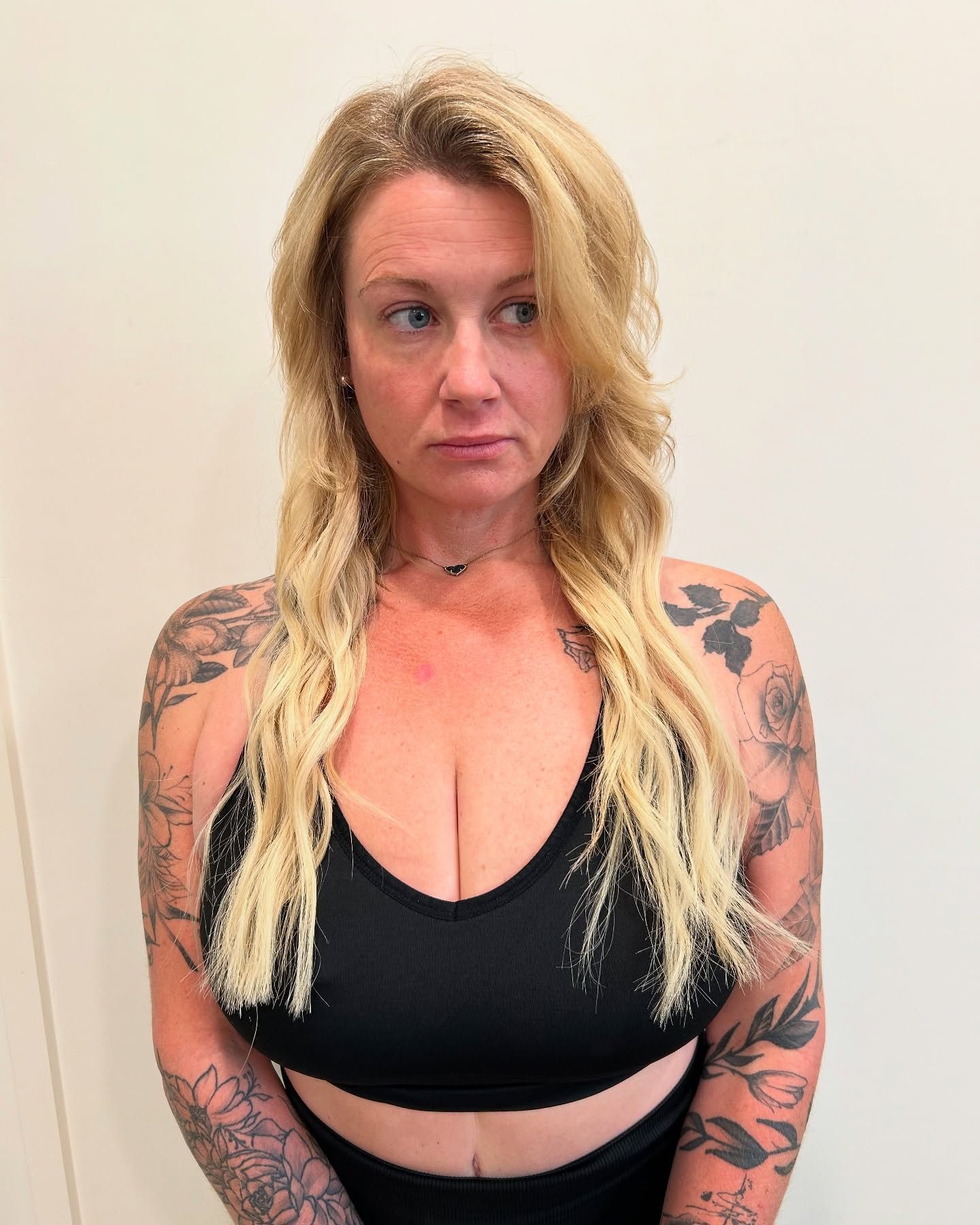Woman with blonde wavy hair and arm tattoos wearing a black sports bra, looking to the side.
