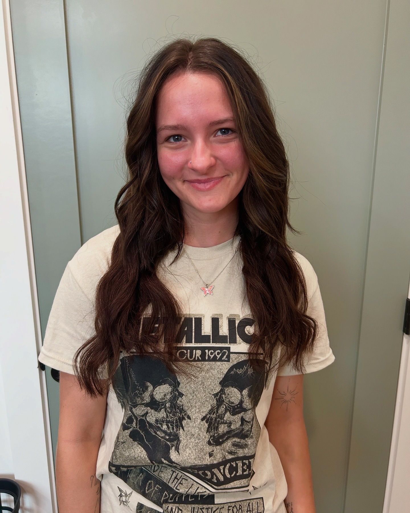 Woman with wavy brown hair wearing a Metallica t-shirt, smiling, standing in front of a neutral-colored wall.