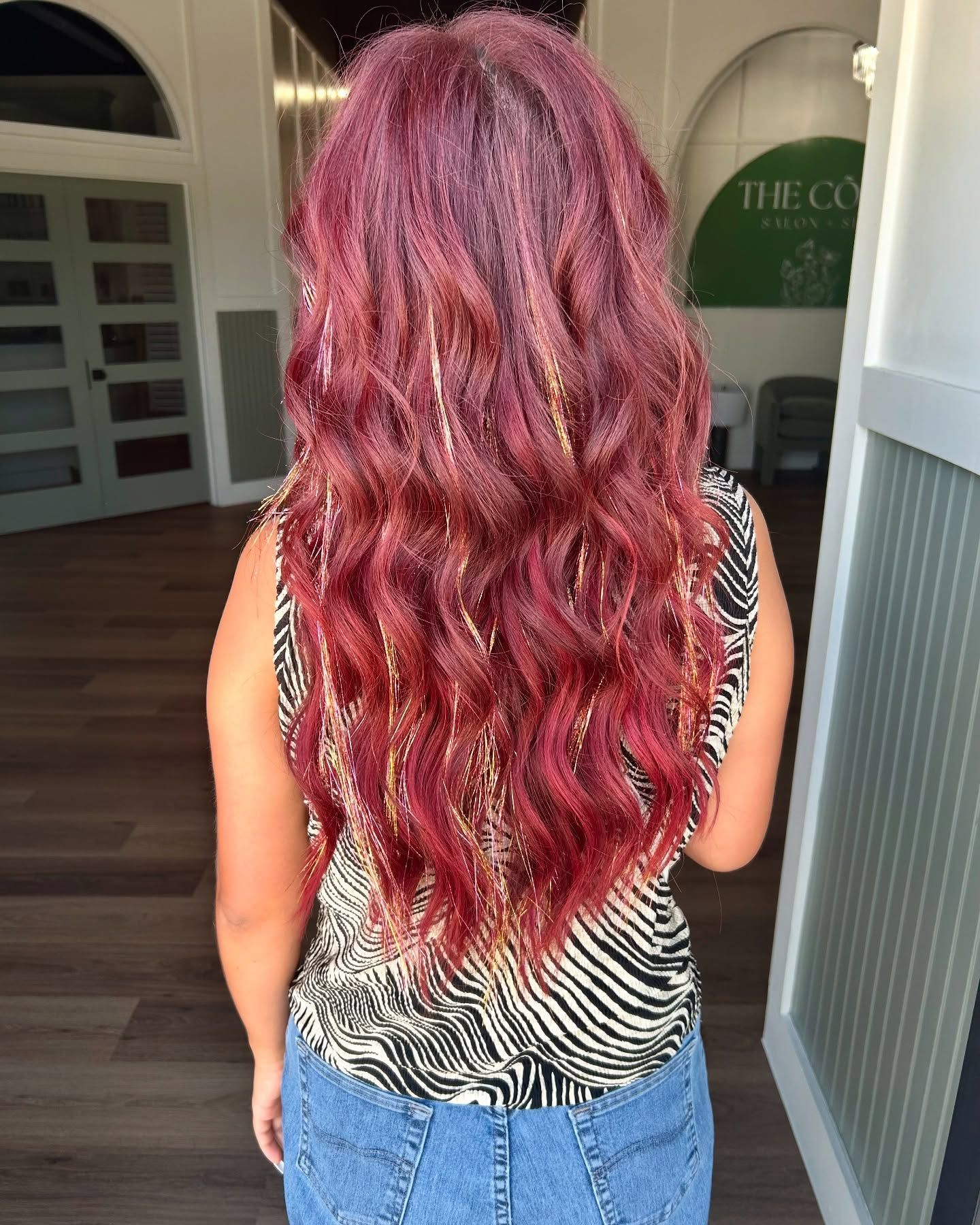 Woman with long, wavy, red hair with gold tinsel. Wearing a zebra print top and jeans. Standing near a door.