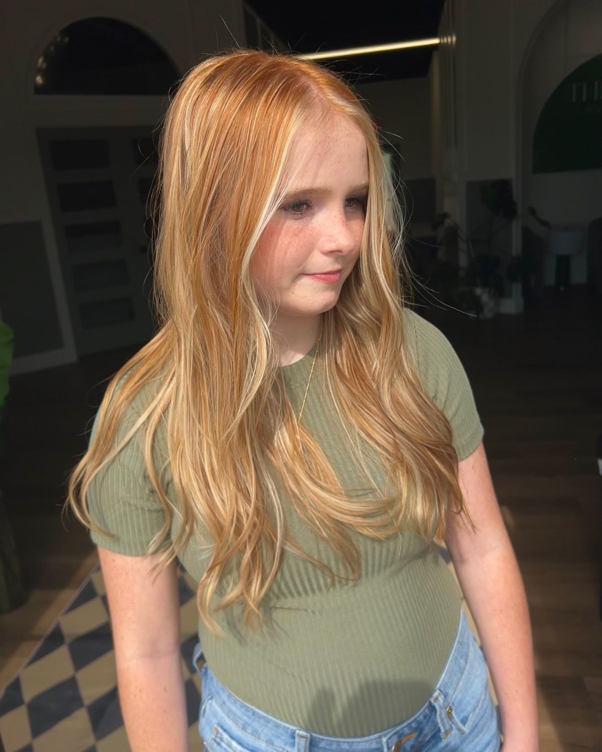 Girl with long wavy hair, wearing a green shirt and denim shorts, stands indoors.
