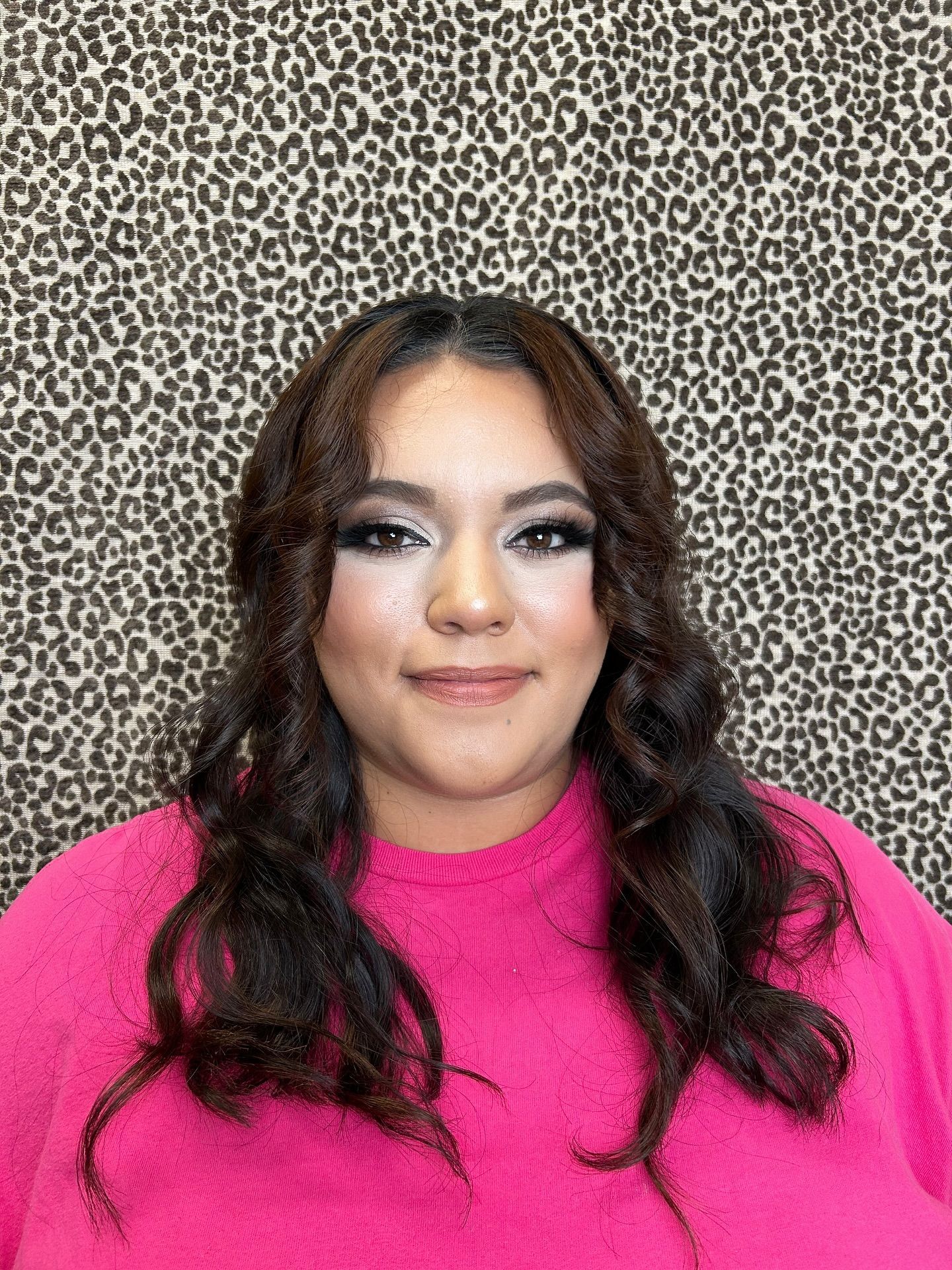 Woman with dark hair, wavy hairstyle, and eye makeup; wearing a pink shirt, in front of a leopard print background.
