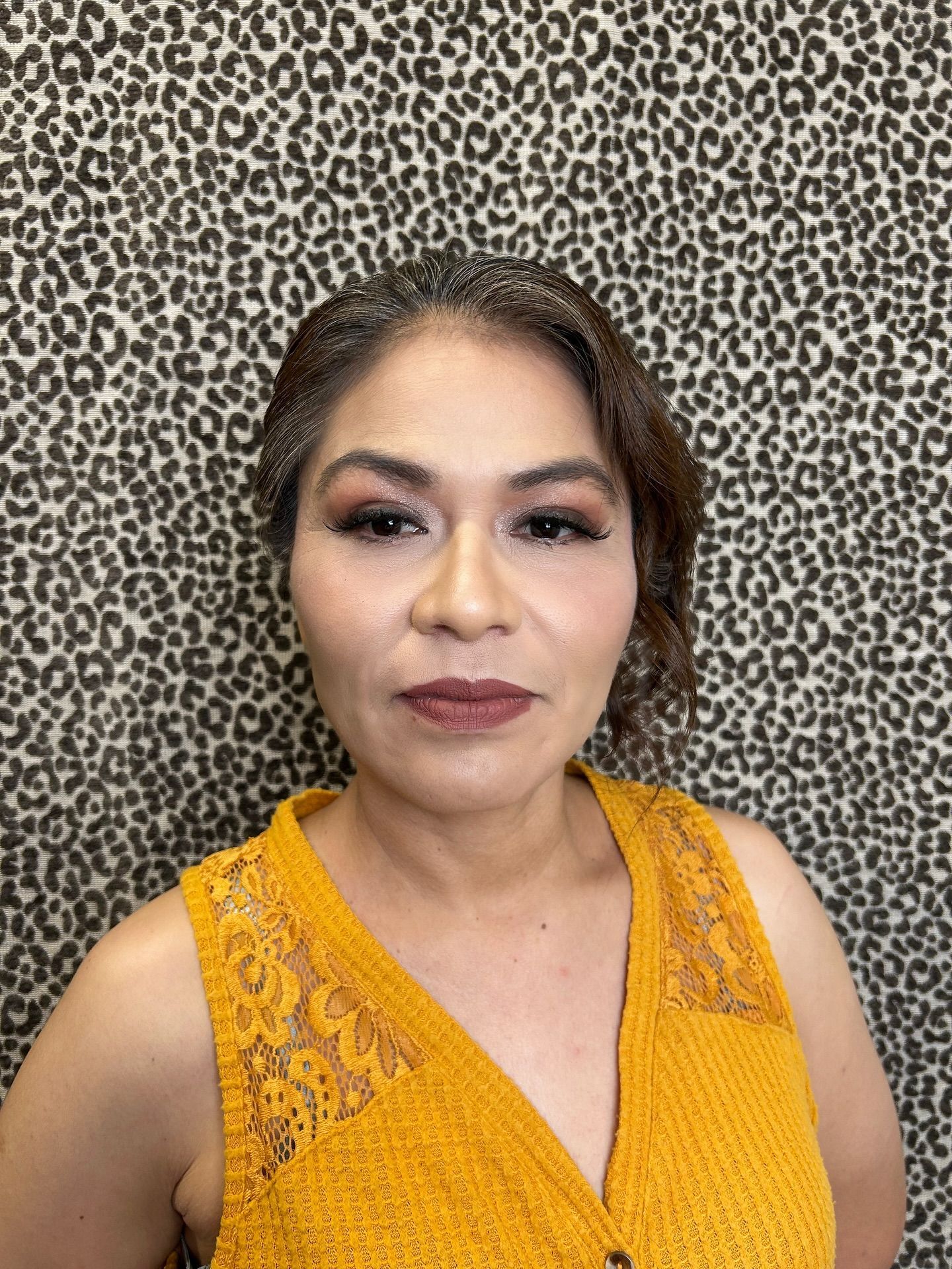 Woman with dark hair and makeup wearing a yellow top against a patterned background.