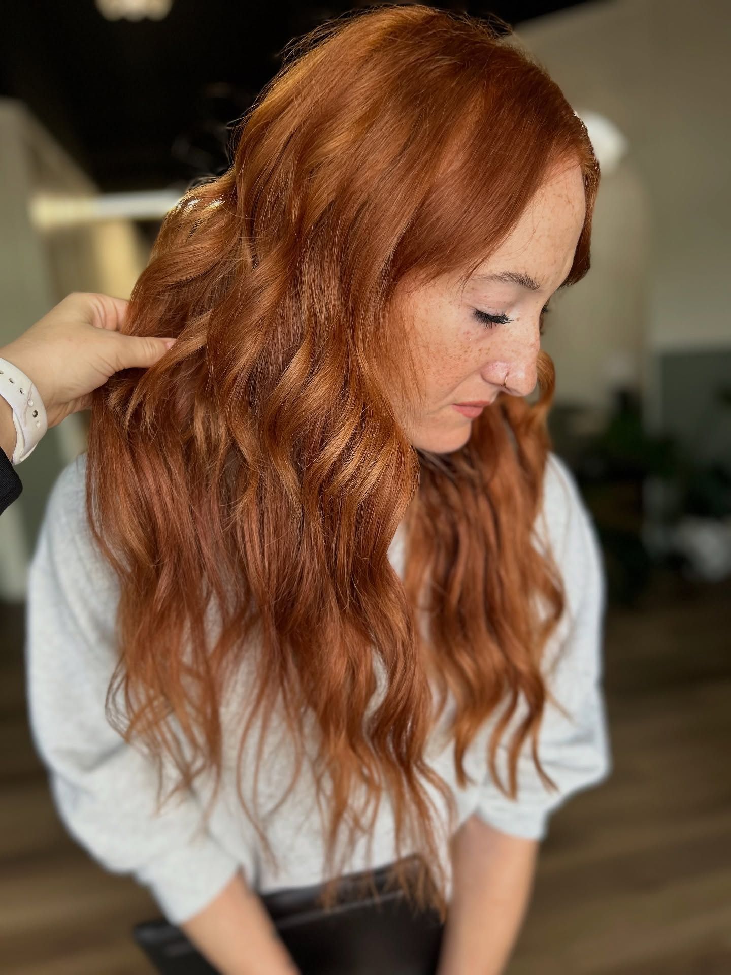 Woman with long, wavy auburn hair. Hand touches hair. Soft, indoor lighting.
