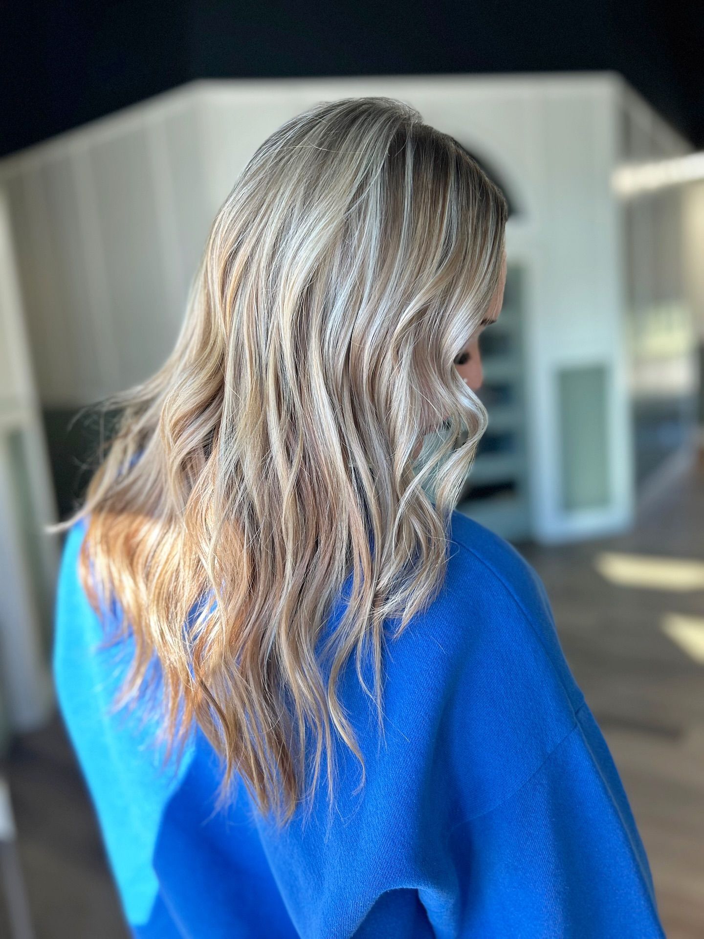 Woman with wavy, light brown hair, wearing a blue sweater, in front of a white building.