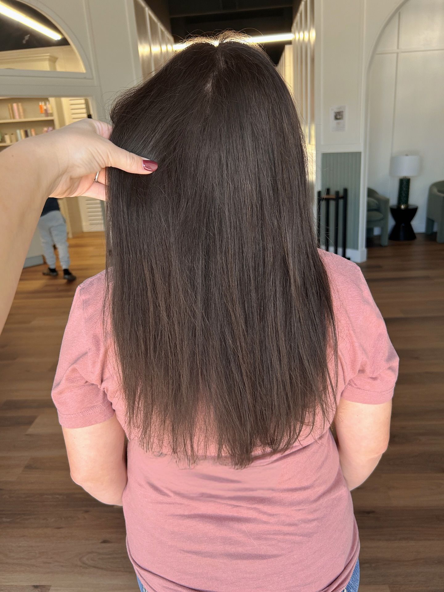 Person's dark, straightened hair. Pink shirt. Hand touching hair. Indoors: wood floors, white walls.