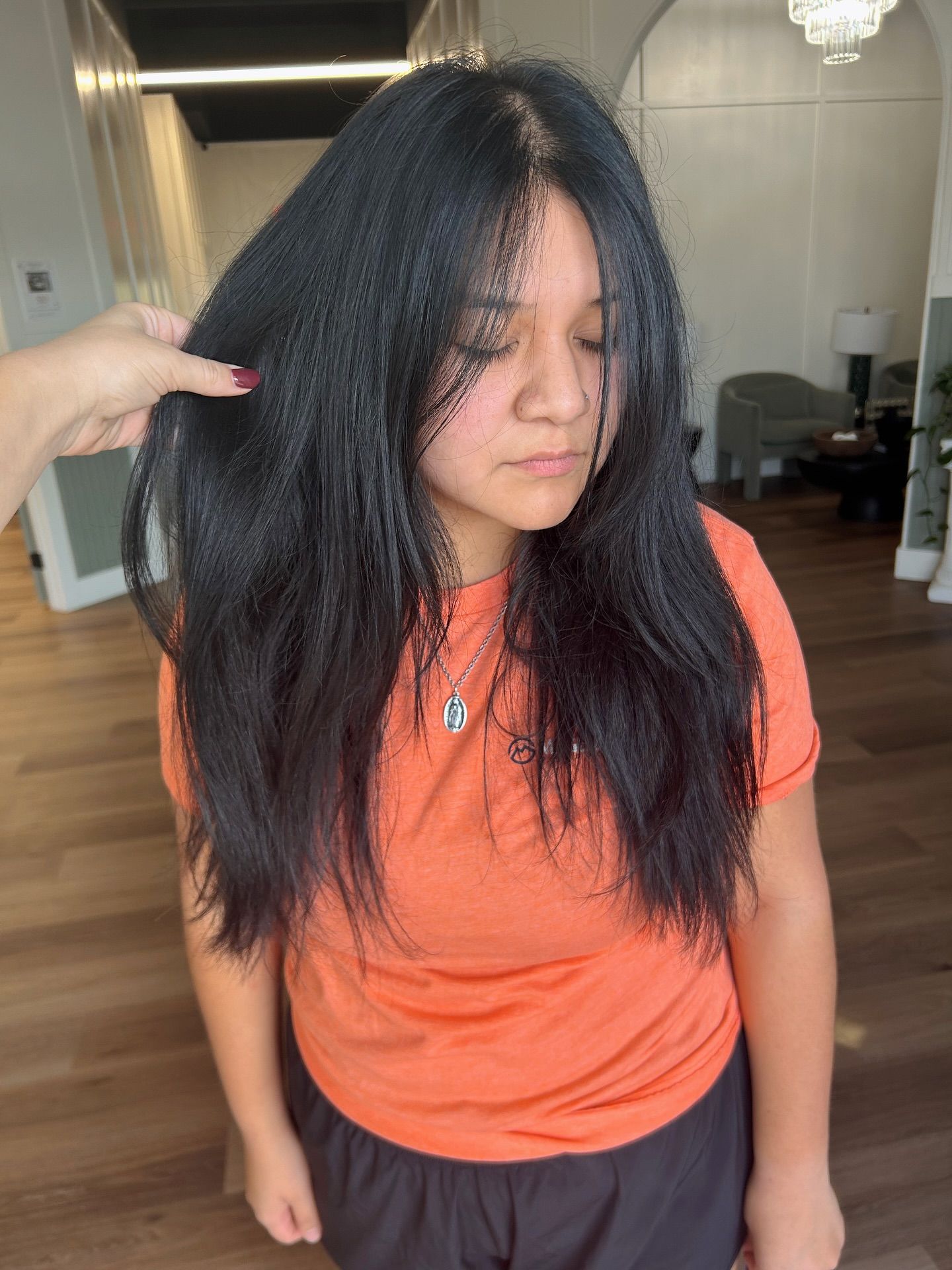 Woman with long black hair, wearing an orange shirt, is having her hair styled in a salon.