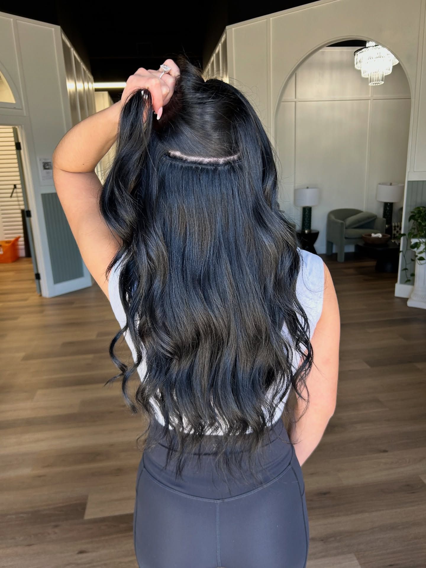 Woman with long black wavy hair, holding top portion up, showing sewn-in extensions.