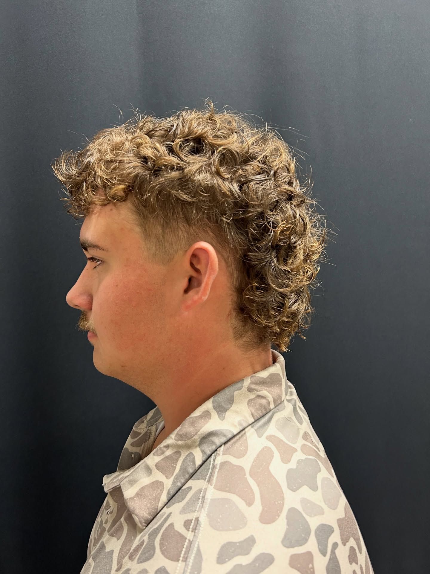 Man with curly brown hair in a mullet style, wearing a patterned button-up shirt, against a dark background.