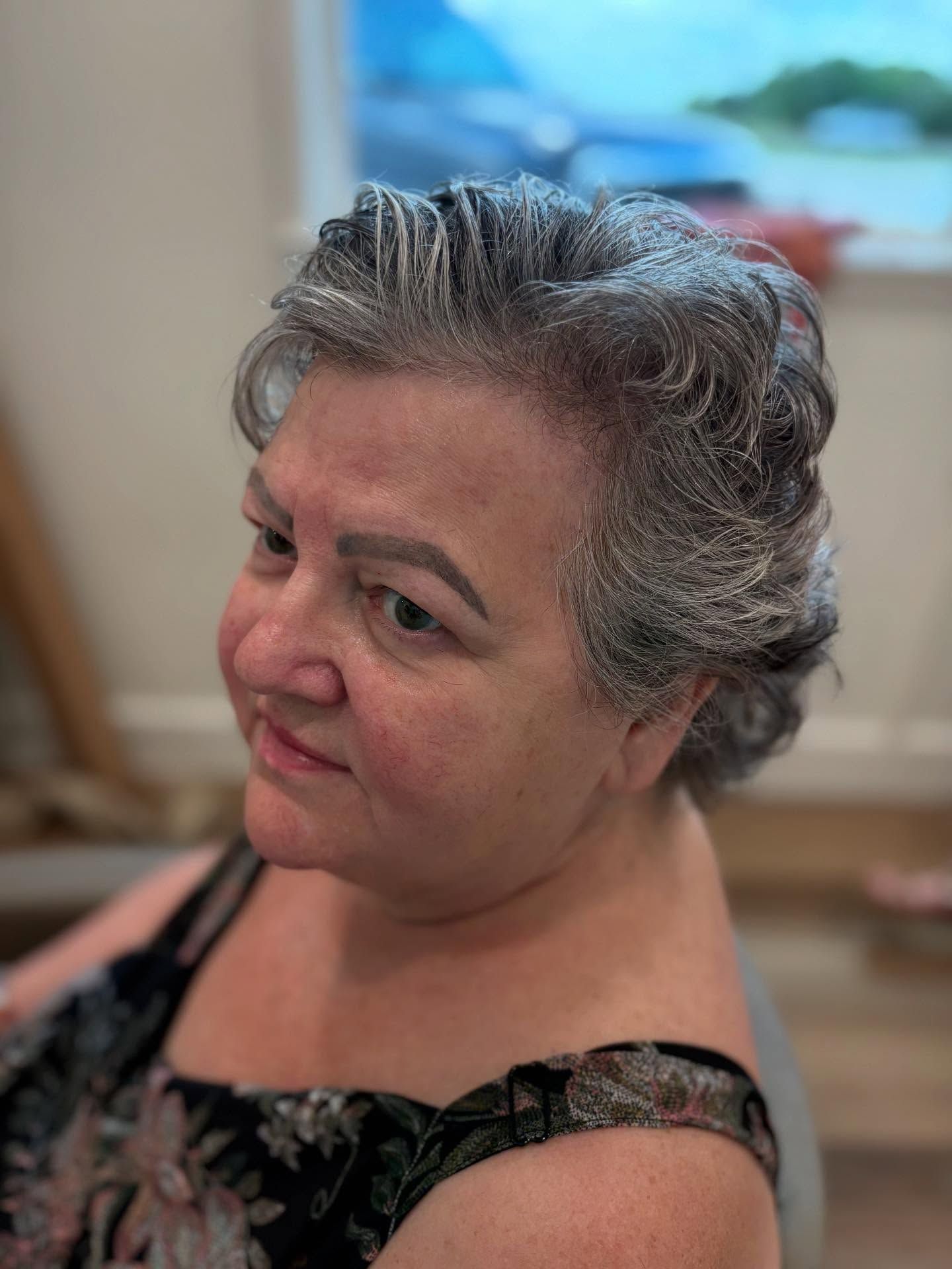 Woman with gray, styled hair looks slightly to the side; indoors with window in the background.