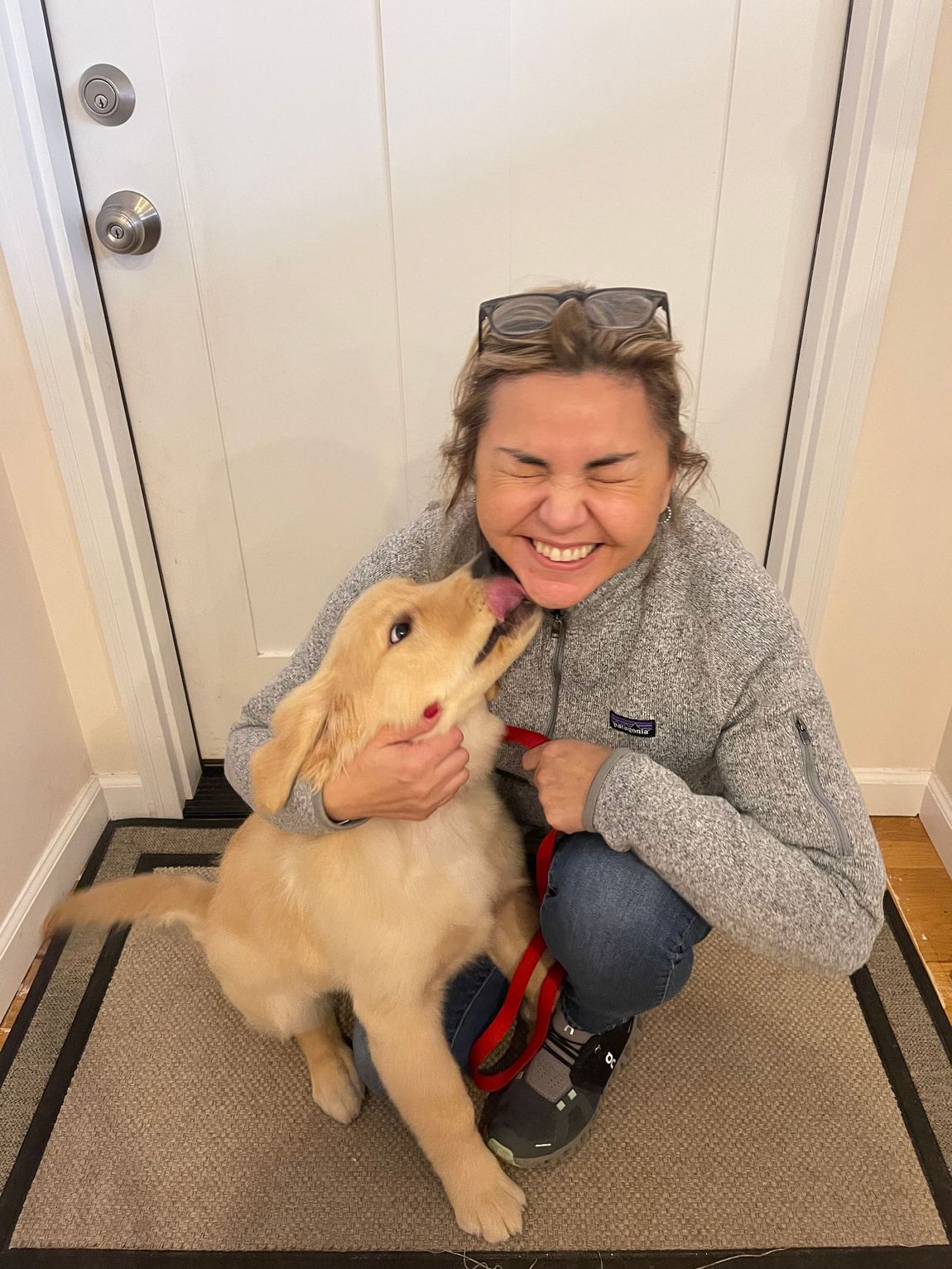 Woman smiles as golden retriever puppy licks her face near a doorway.
