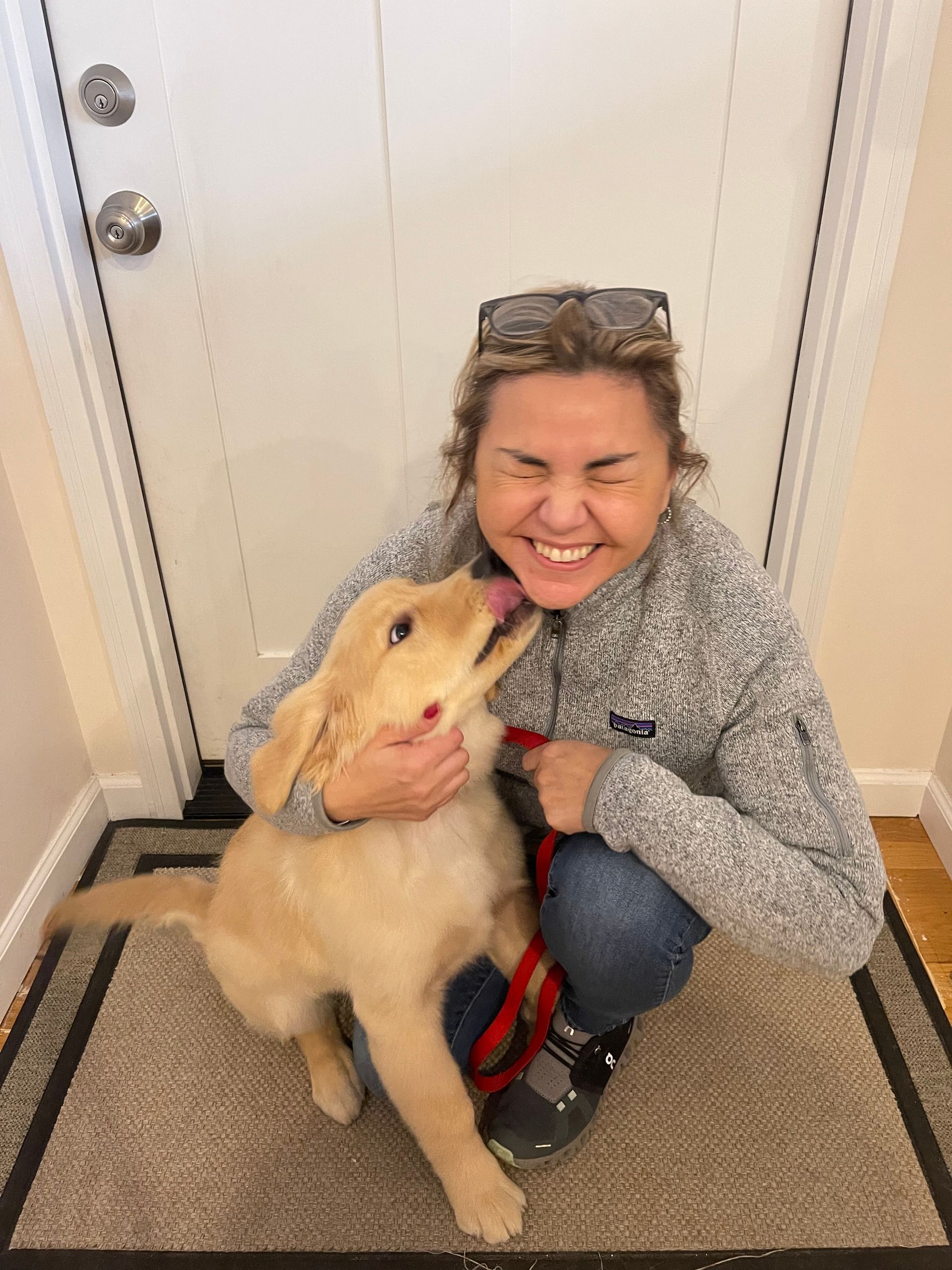 Woman smiles as golden retriever puppy licks her face near a doorway.