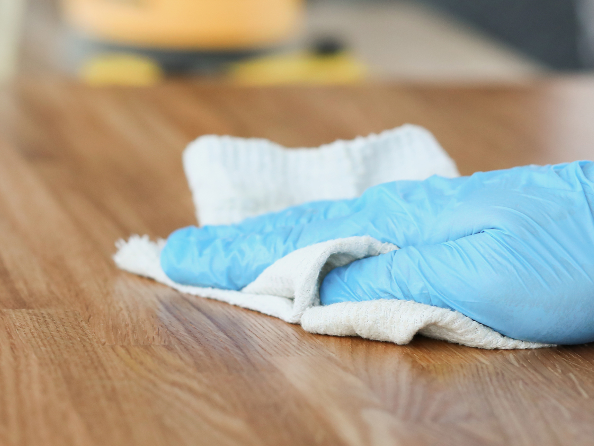 Blue-gloved hand wiping a wooden table with a white cloth