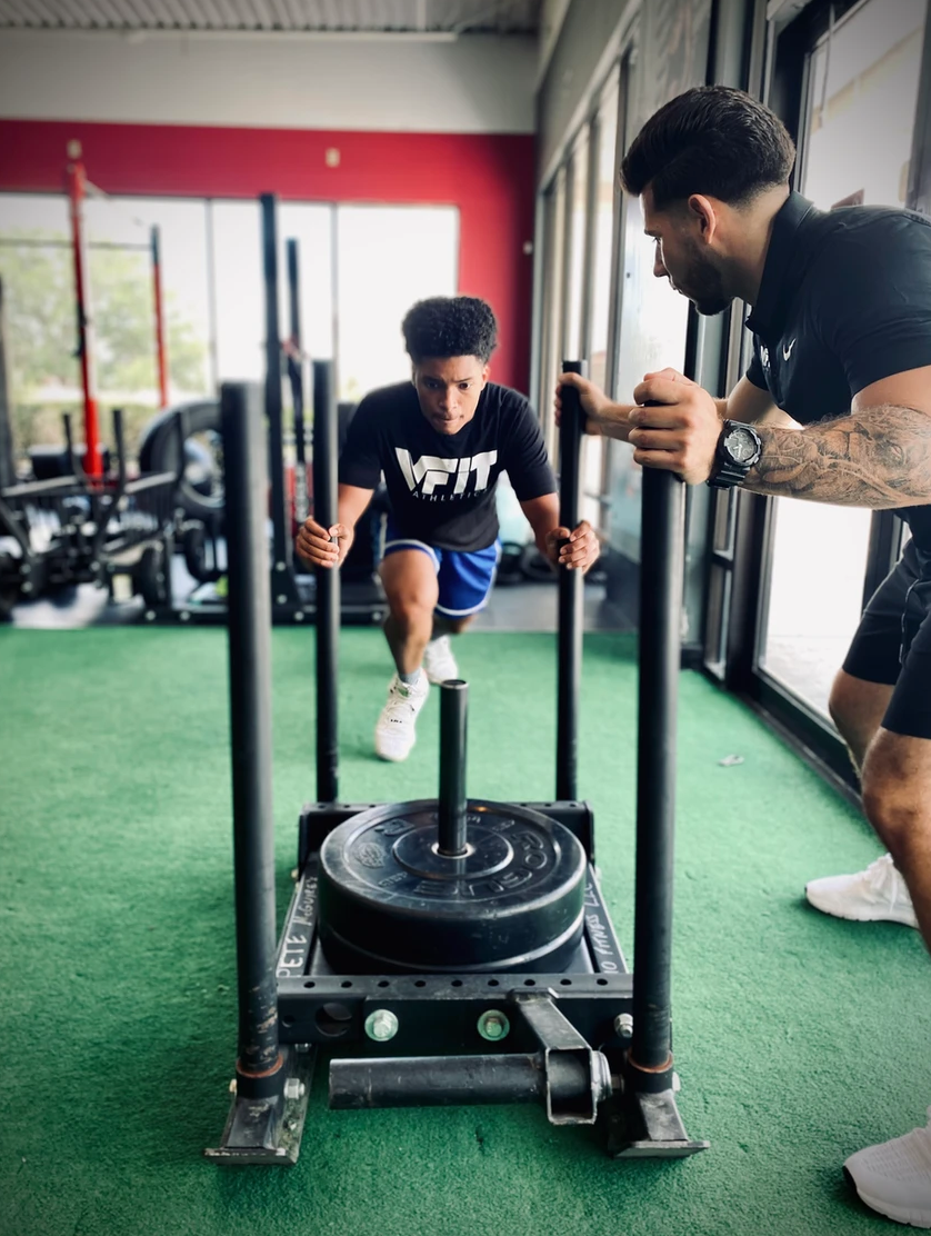 A man is pushing a sled in a gym while another man watches.