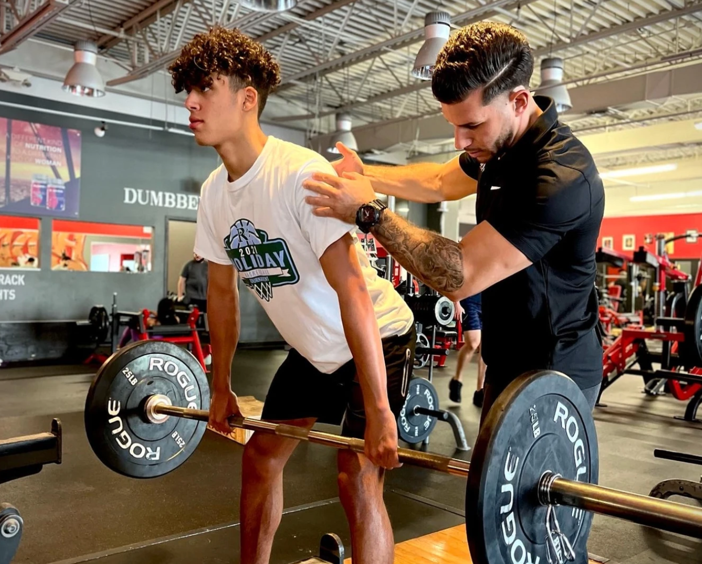 A man is helping a young man lift a barbell in a gym.